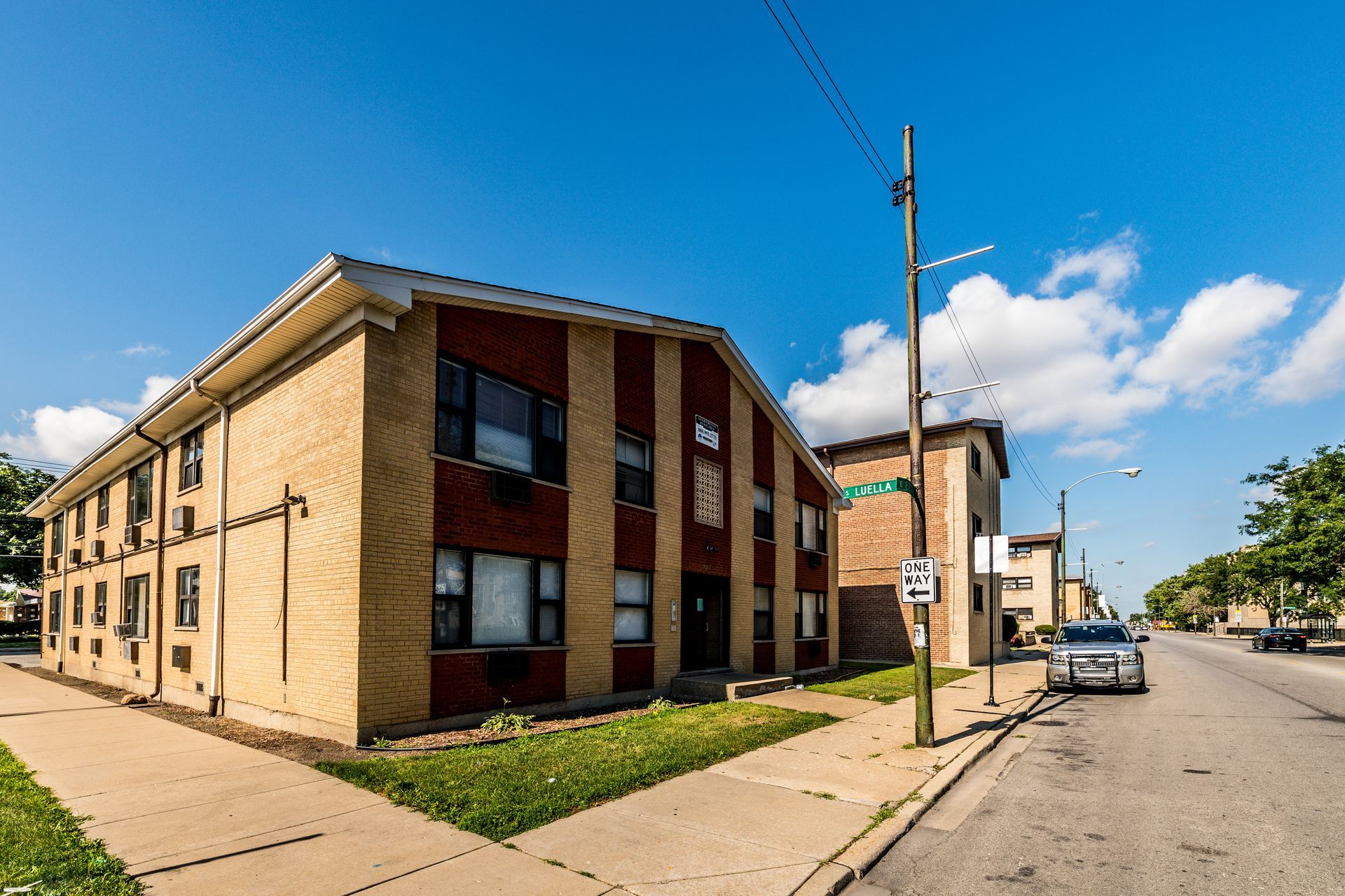 Two-story brick apartment building on a sunny street, with grass and parked cars visible.