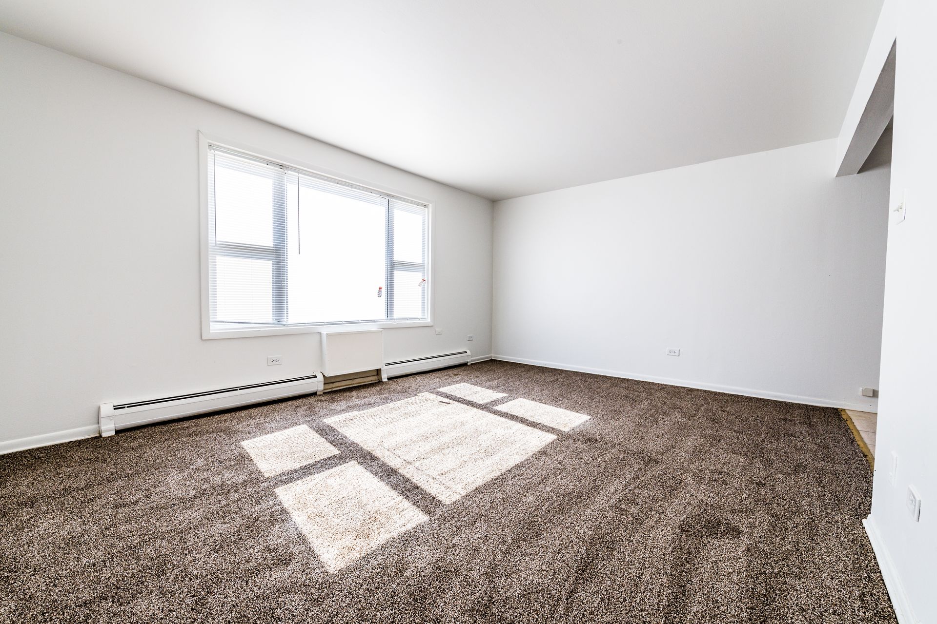 Empty living room with brown carpet, large window, and white walls.