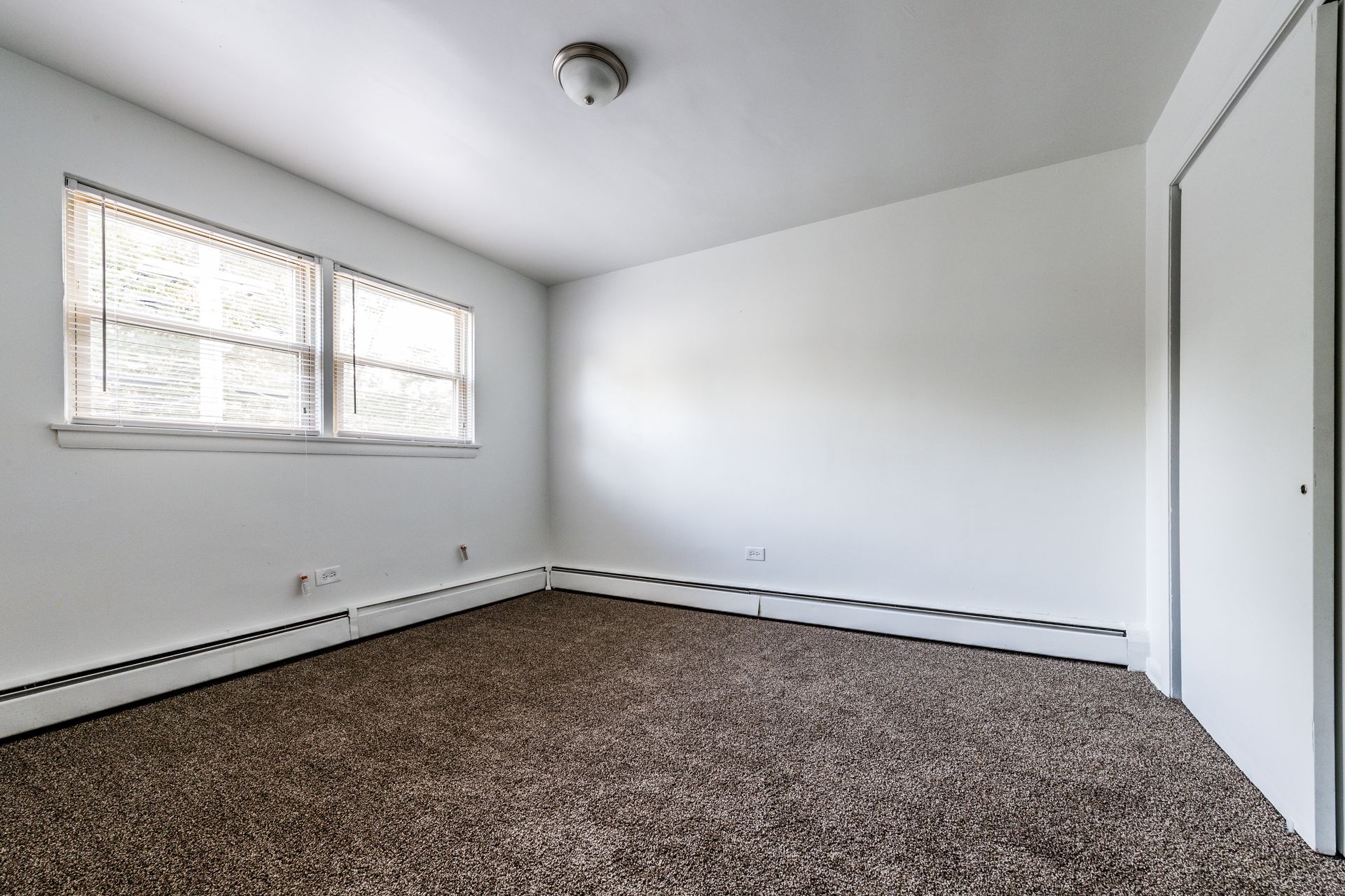 Empty room with brown carpet, white walls, small window with blinds, and a closed door.