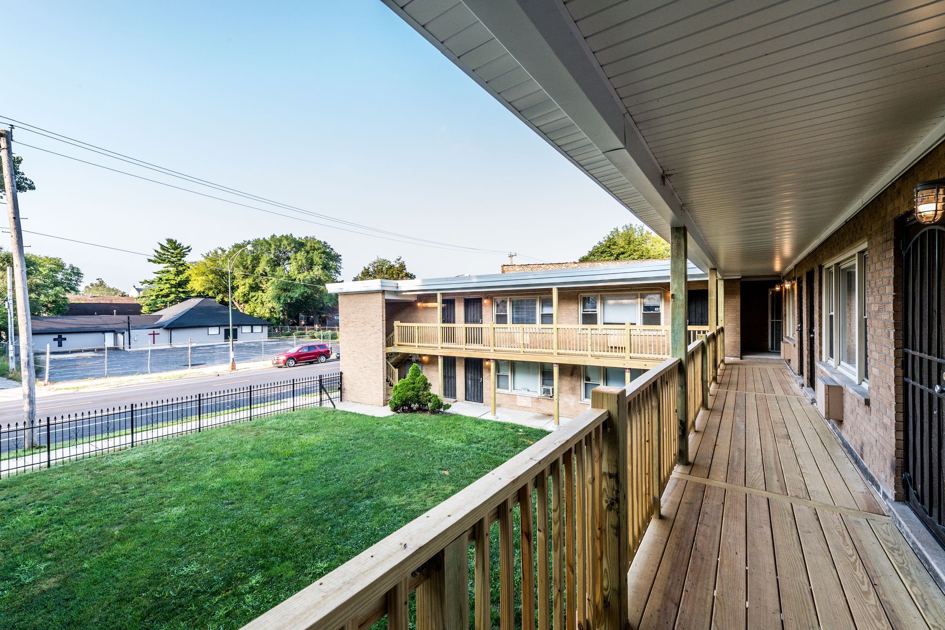 Balcony view of two-story brick apartment building and street. Green grass and fence in foreground, blue sky.