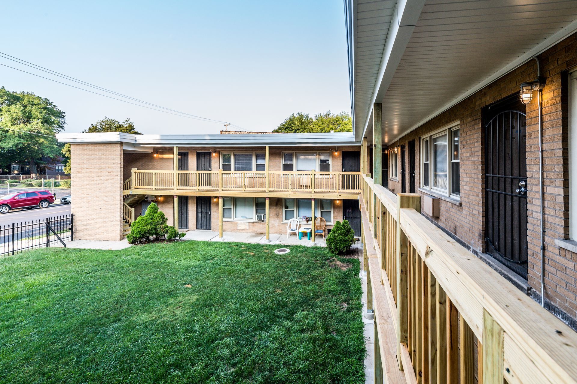 Two-story brick apartment building with wooden balconies. Green lawn in the foreground, street and parked car on the left.