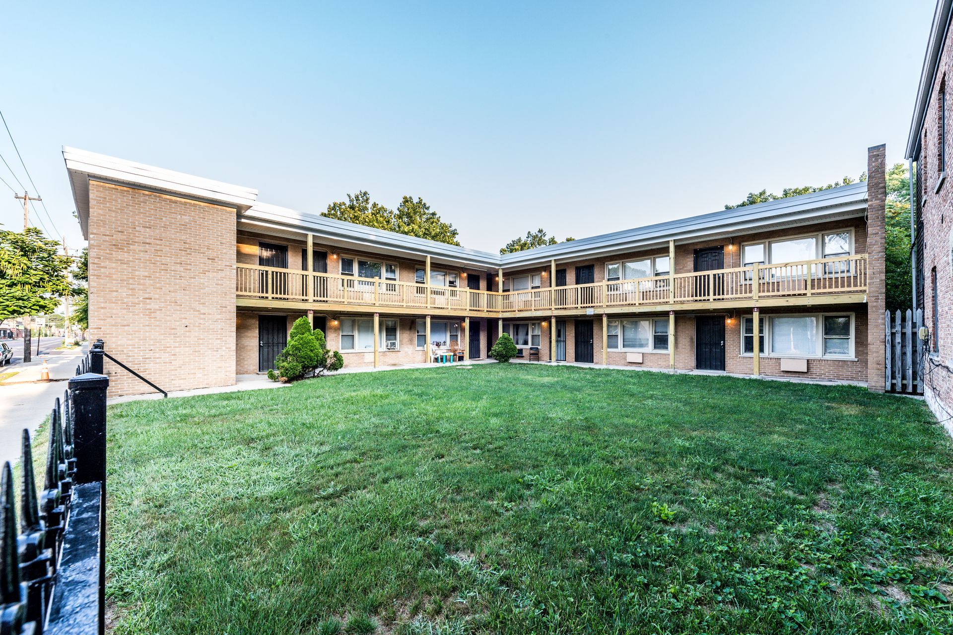 Two-story brick apartment building with wooden balconies, a green lawn, and a black fence in front.