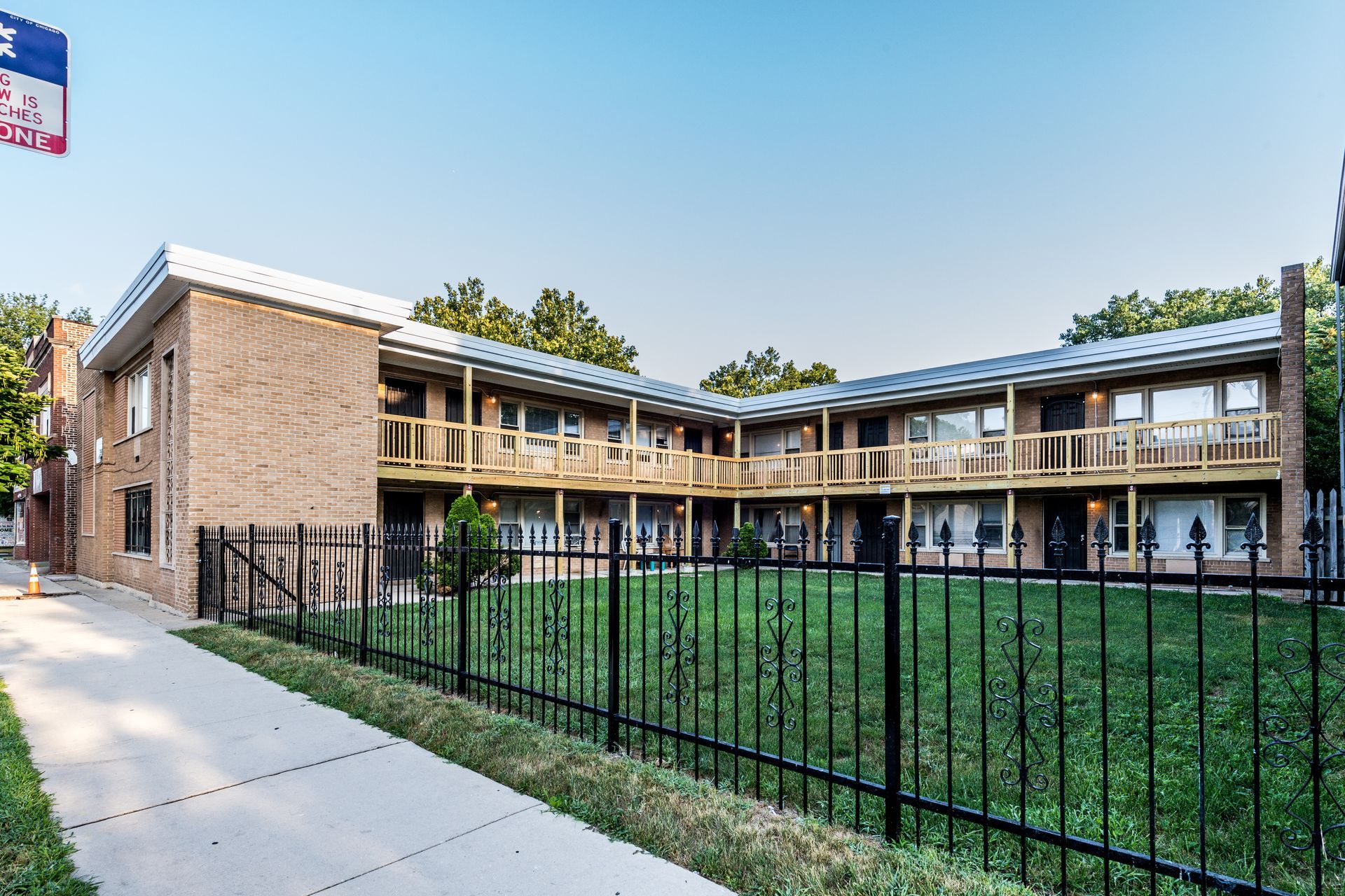 Two-story brick apartment building with a black fence, green lawn, and sidewalk.