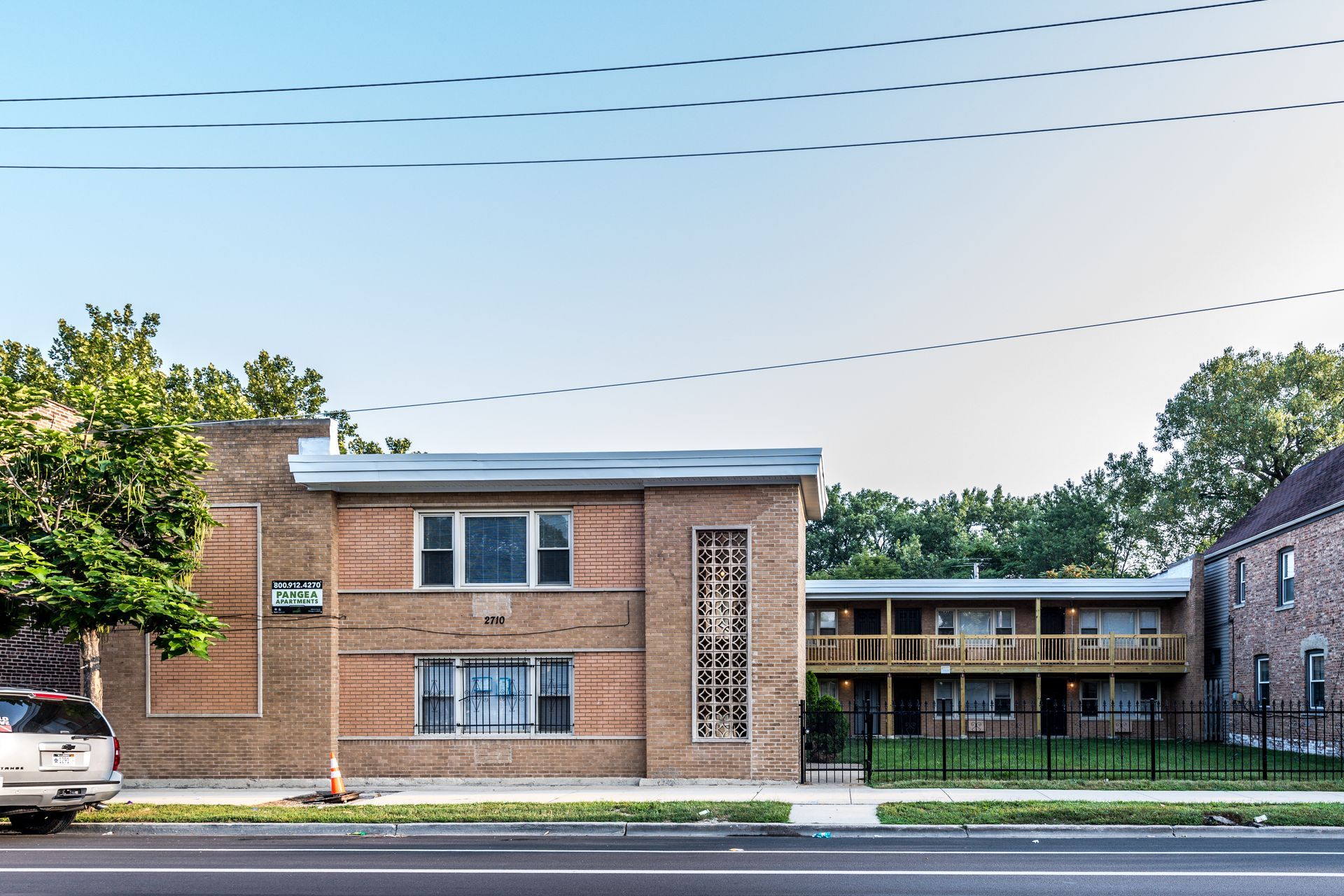 Two-story brick apartment building with a flat roof. Trees and a fence in front of a green lawn and a street.