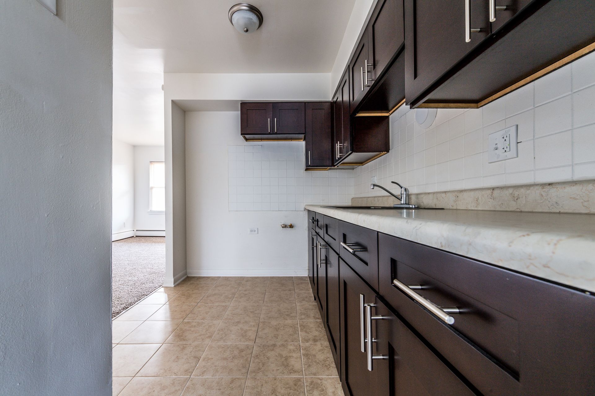 Kitchen with dark wood cabinets, light countertops, and tile backsplash; hallway visible in the background.