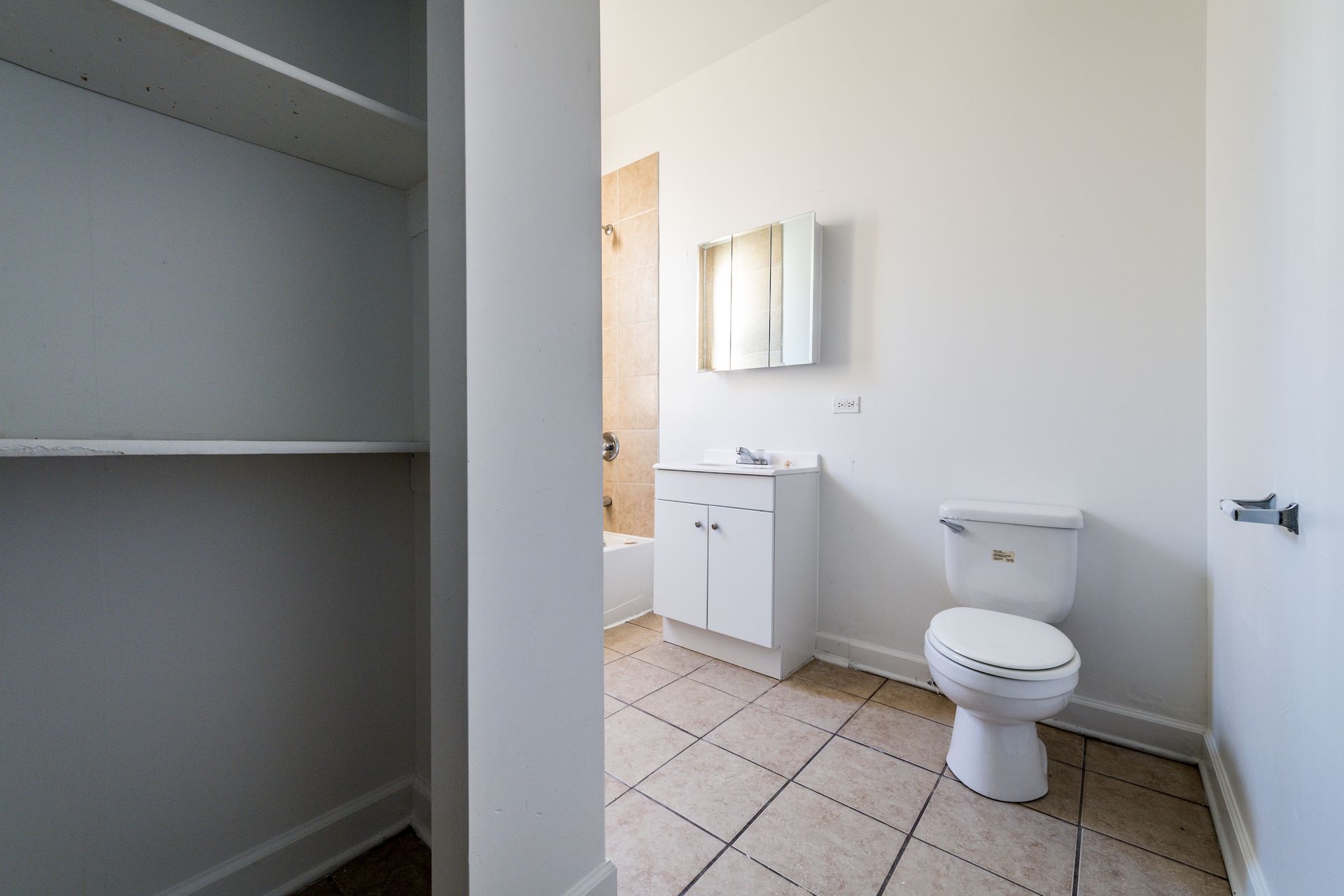 Empty bathroom with white walls, cabinets, toilet, and tan tile floor.