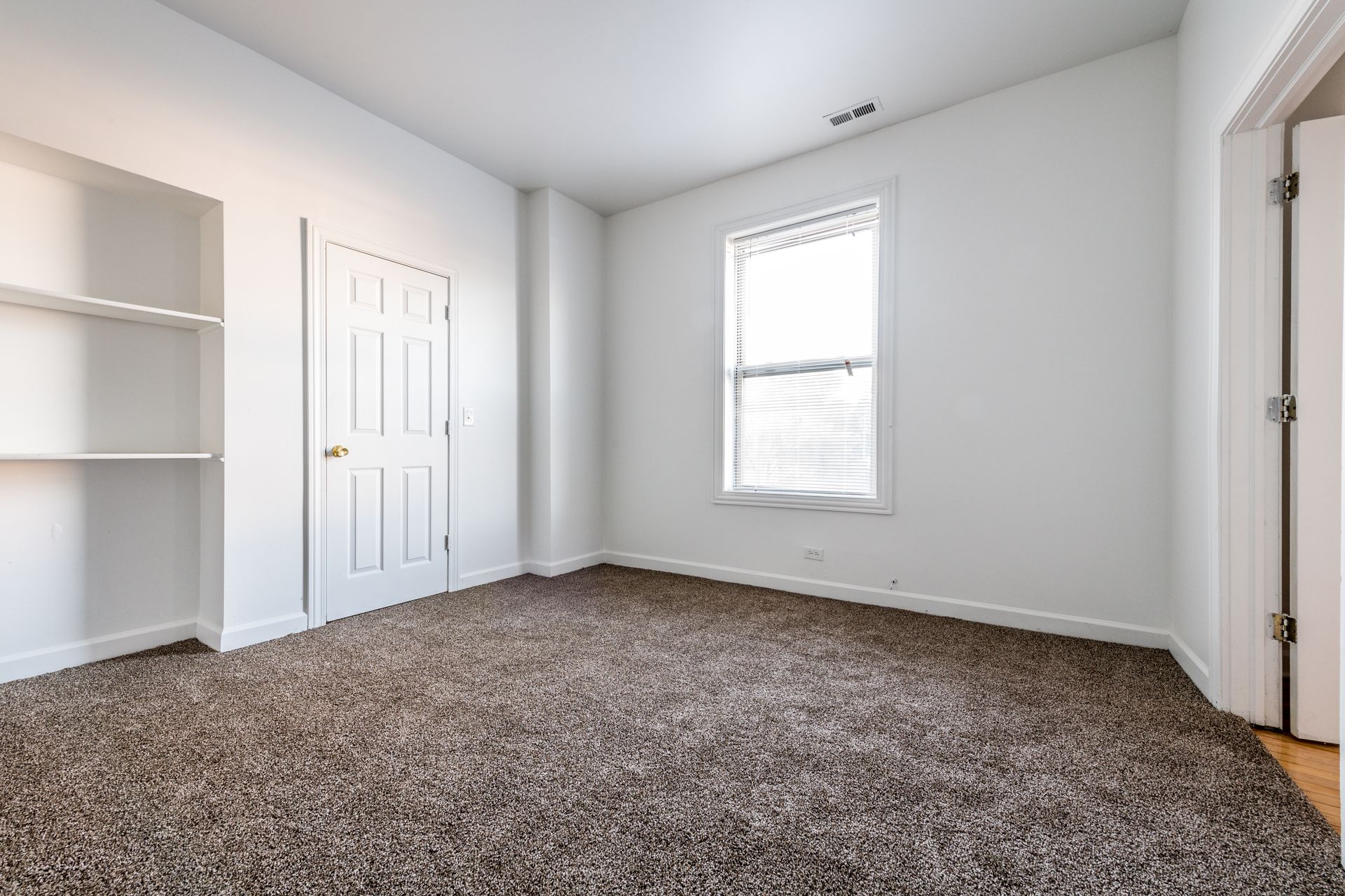 Empty room with brown carpet, white walls, built-in shelves, door, and window.