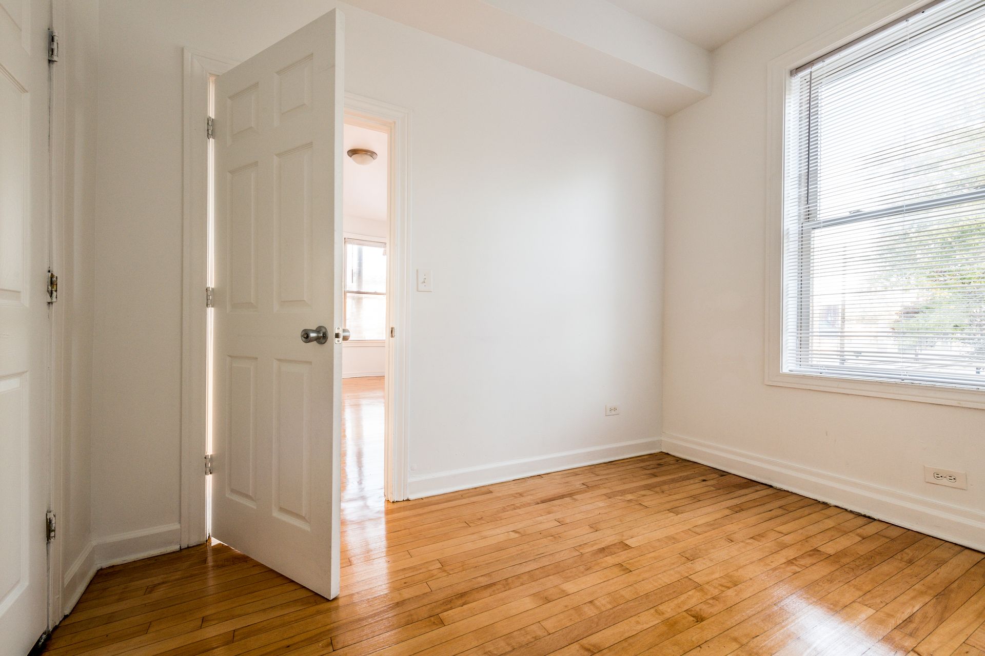 Empty room with hardwood floors, white walls, and a partly open door. A window is on the right.
