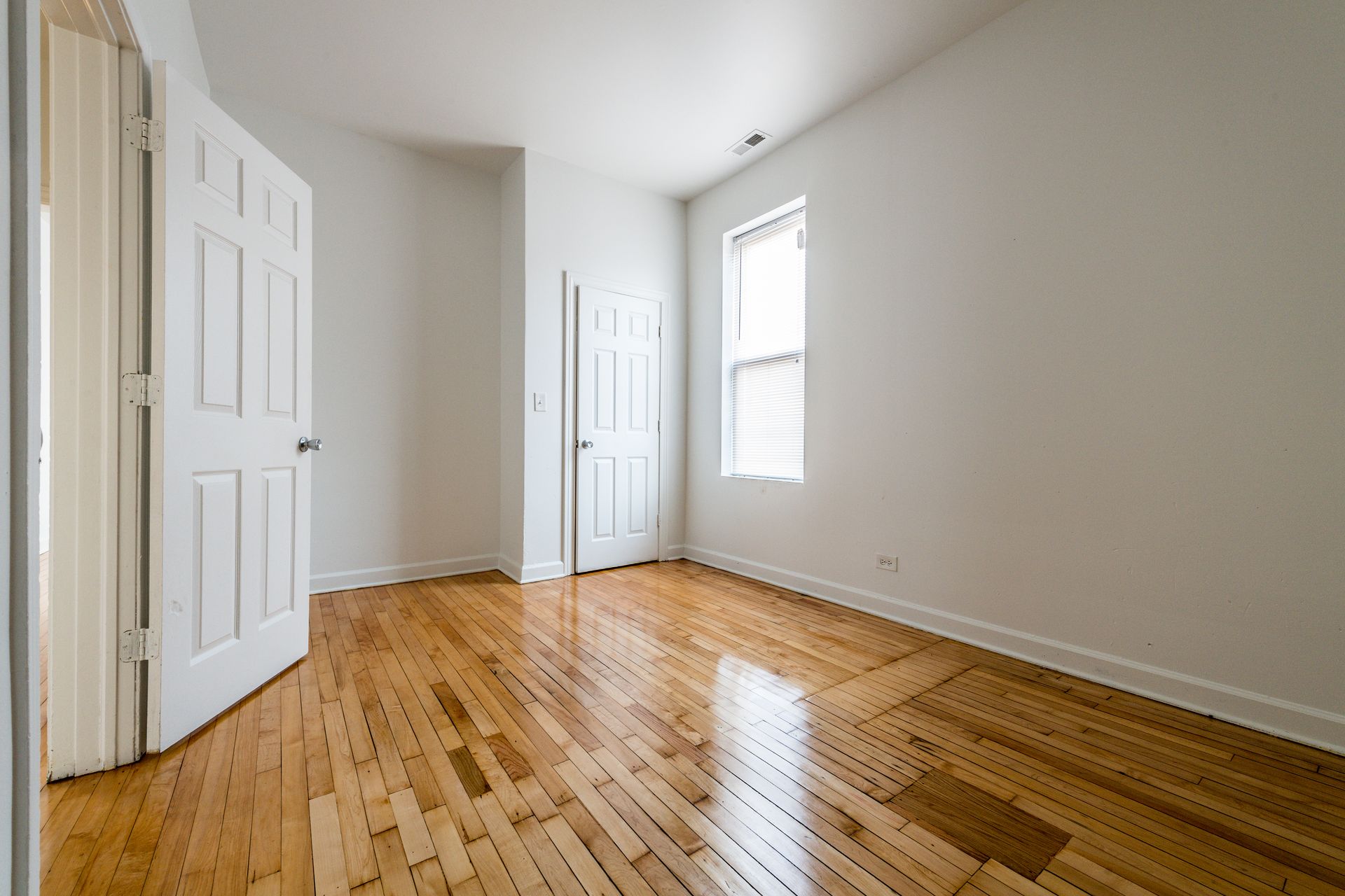 Empty room with hardwood floors, white walls, two doors, and a window.