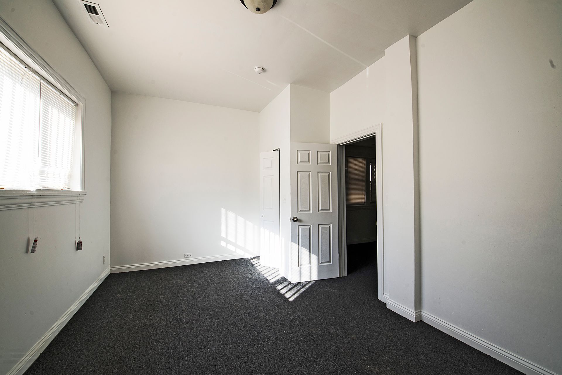 Empty room with white walls, window with sheer curtain, gray carpet, and a doorway.