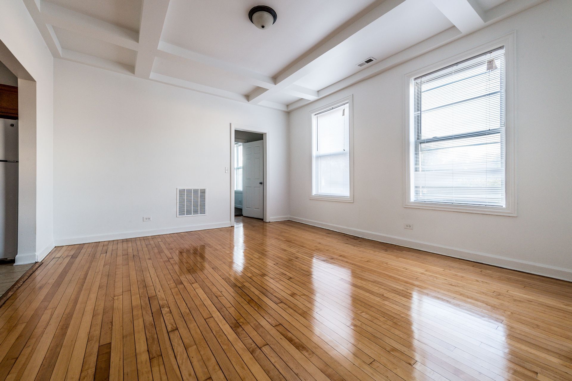 Empty room with hardwood floors, white walls, two windows, and overhead beams.