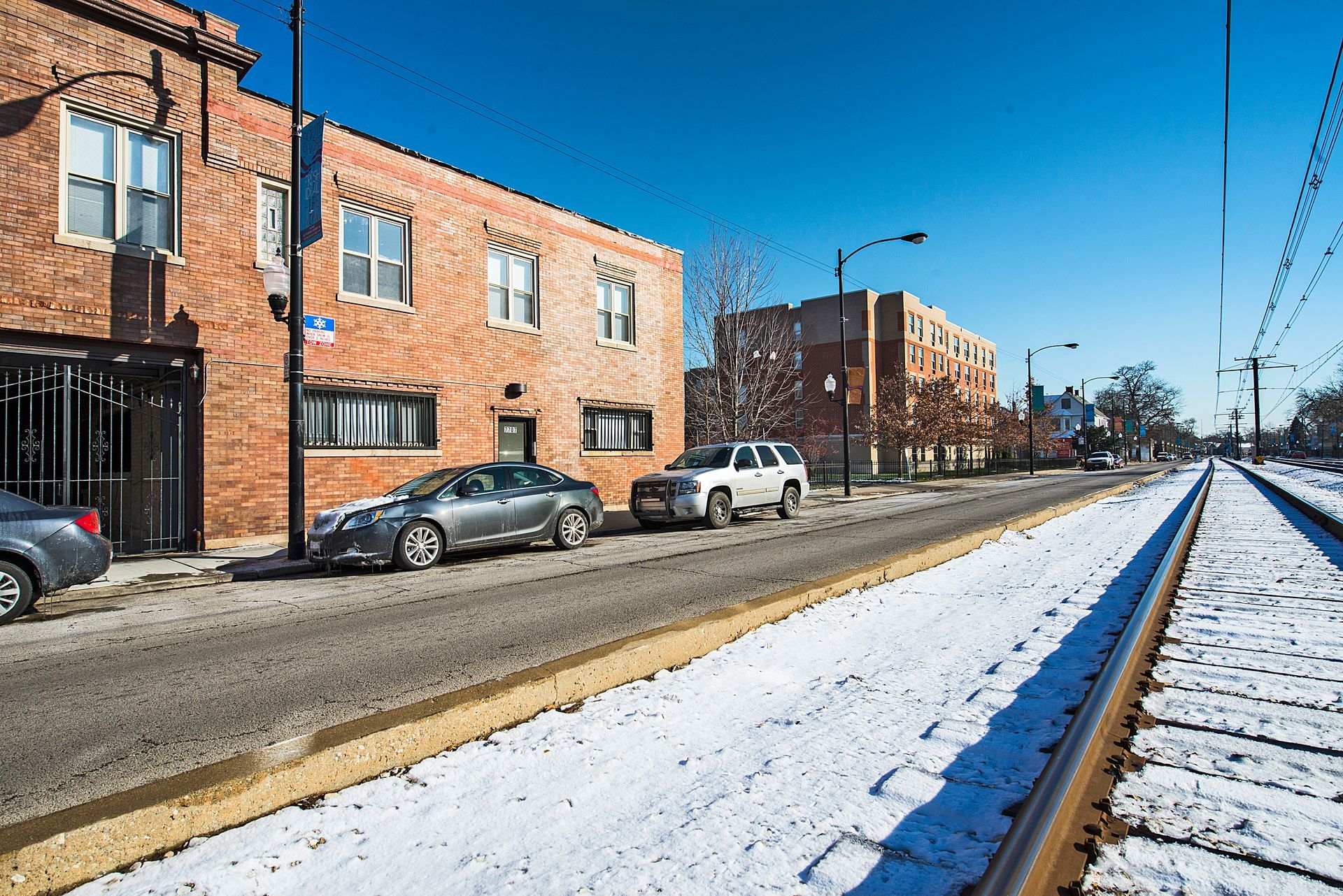Street scene in winter with snow-covered train tracks, brick buildings, parked cars, and a clear blue sky.