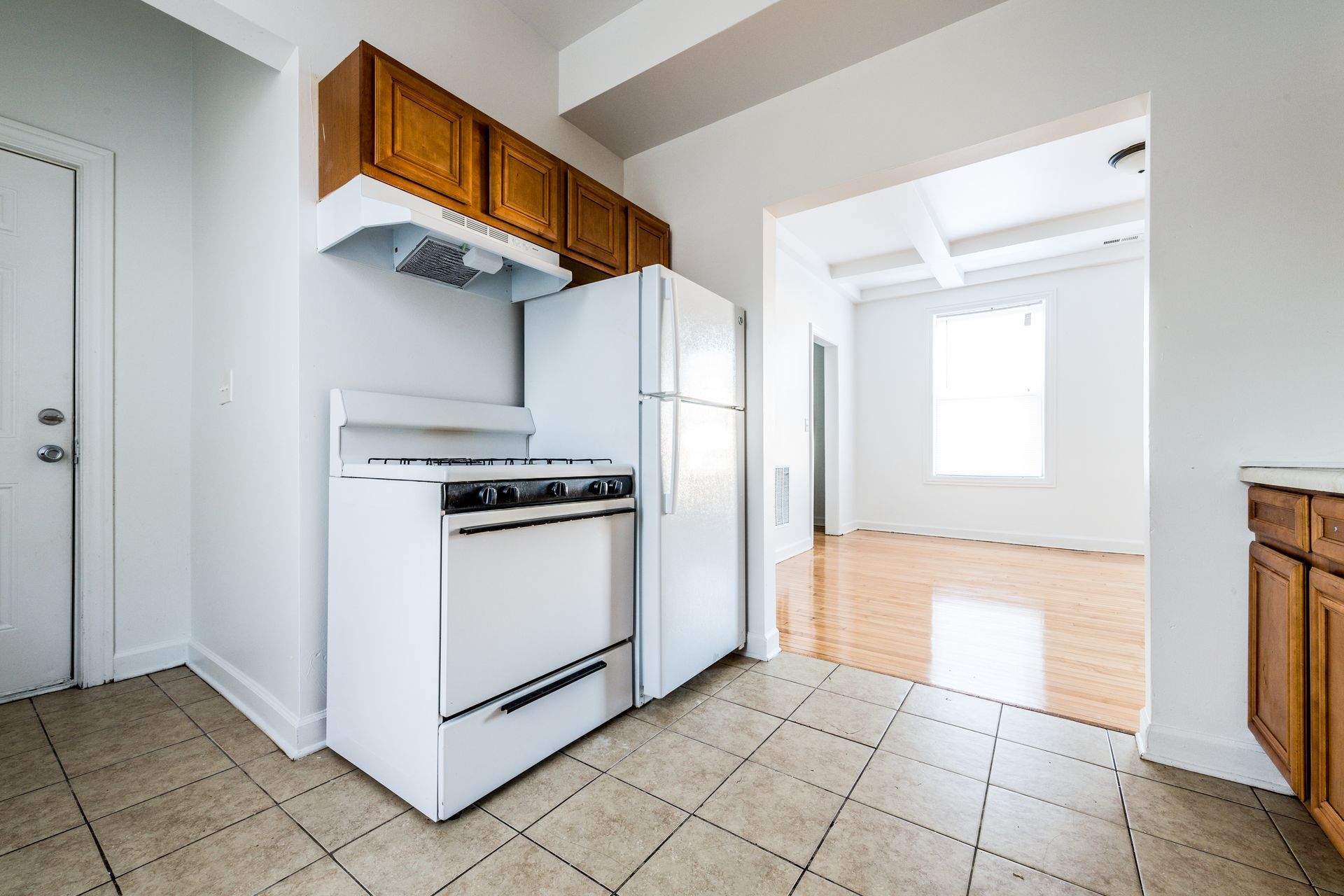 White kitchen with stove, refrigerator, cabinets, doorway to living area, and tile floor.