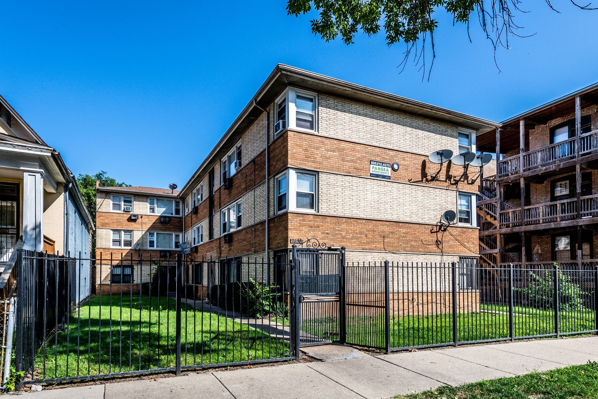 Three-story brick apartment building behind a black fence on a sunny day.