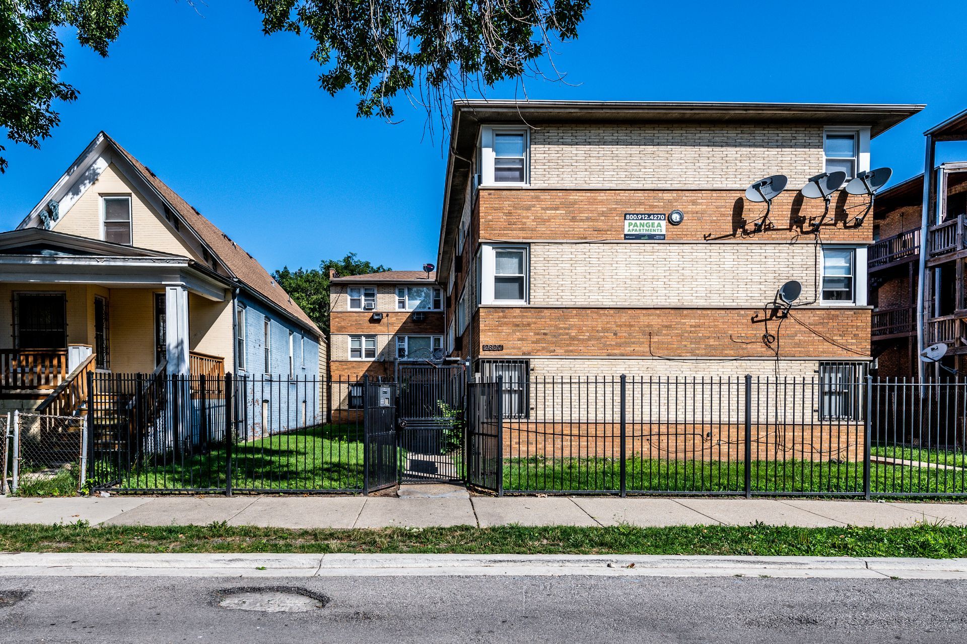 Buildings behind a black metal fence on a sunny day. A two-story brick building and a smaller wooden house are visible.