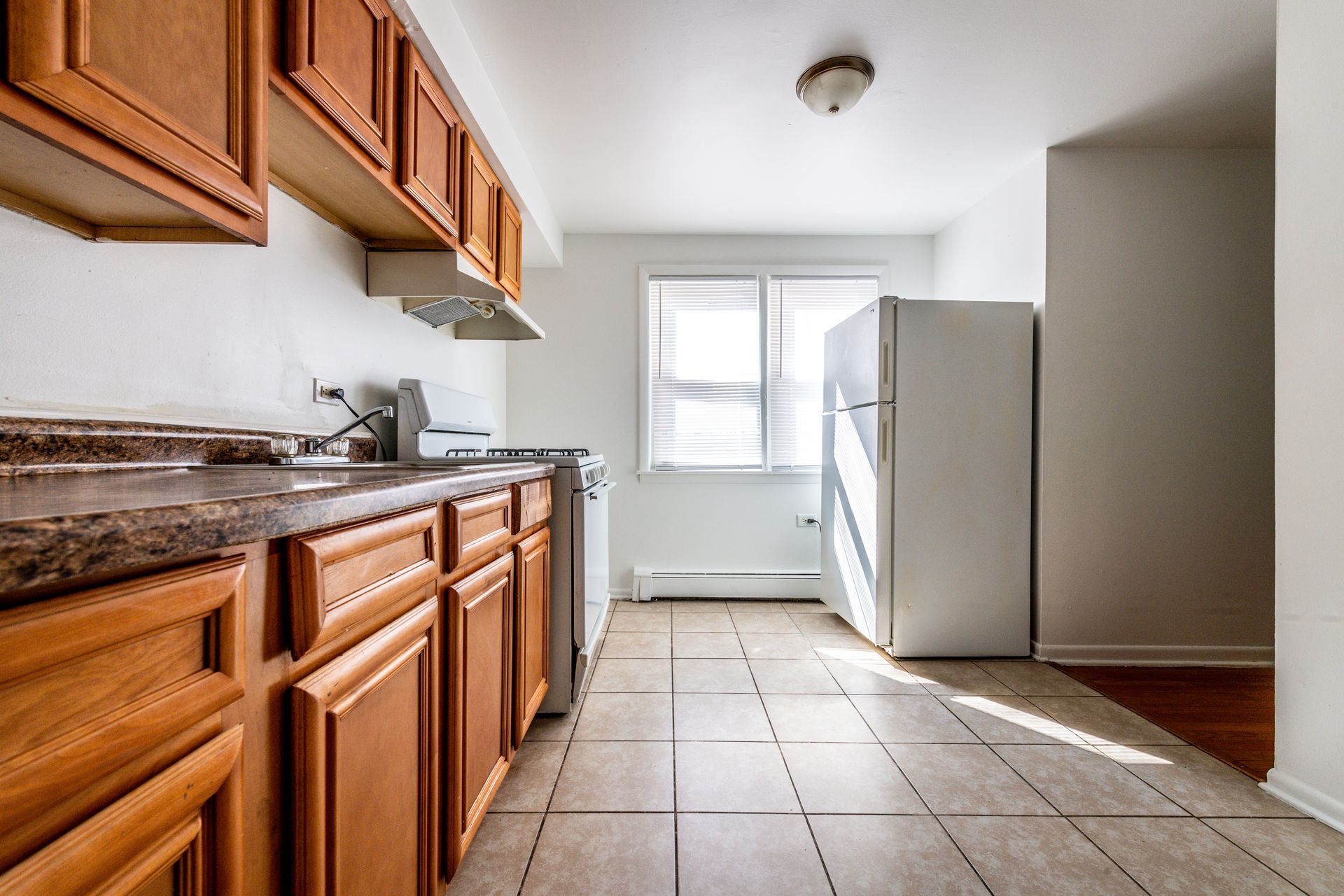 Kitchen with wooden cabinets, beige tiled floor, and a white refrigerator next to a window.