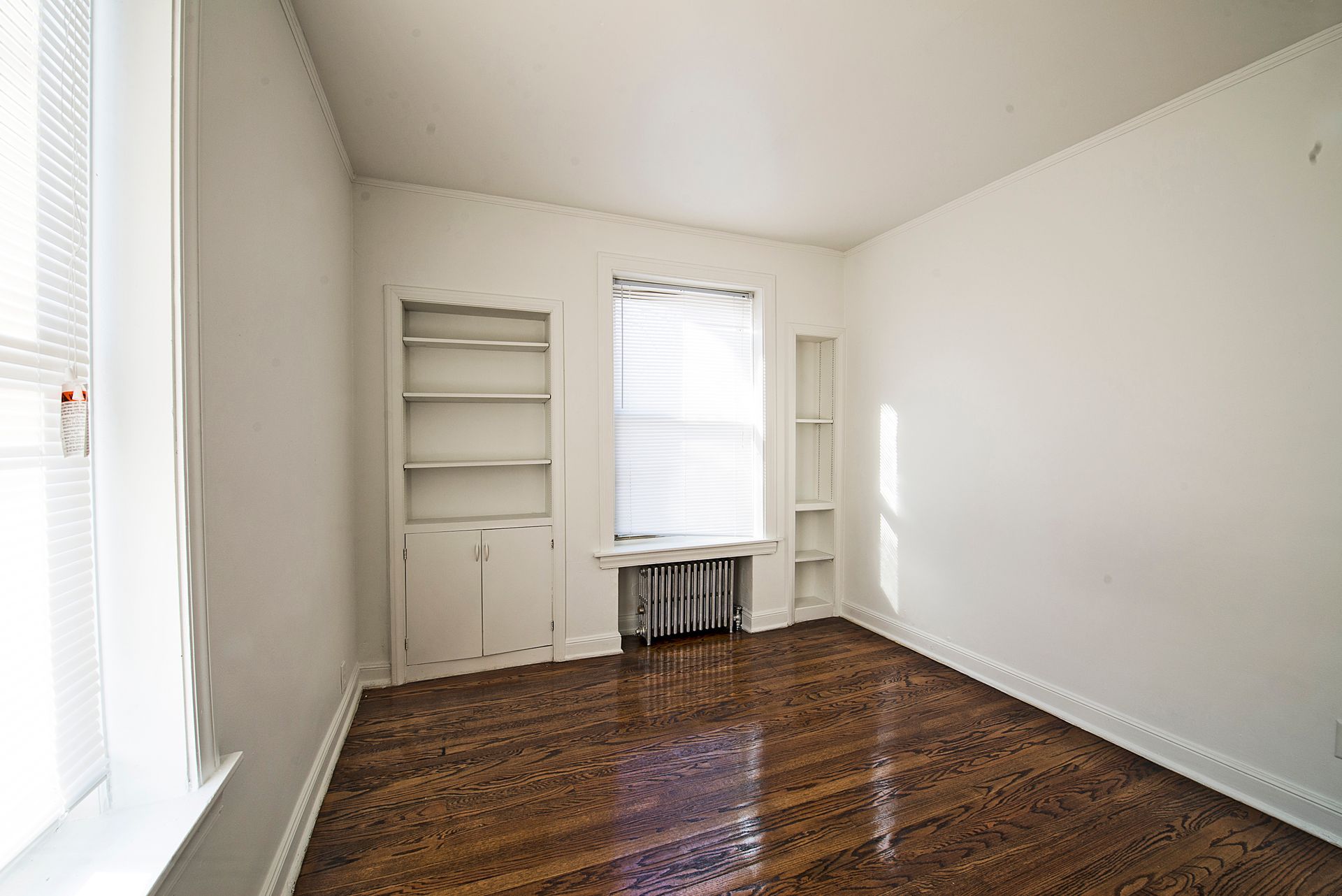 Empty room with hardwood floors, built-in shelving, and a window with blinds.