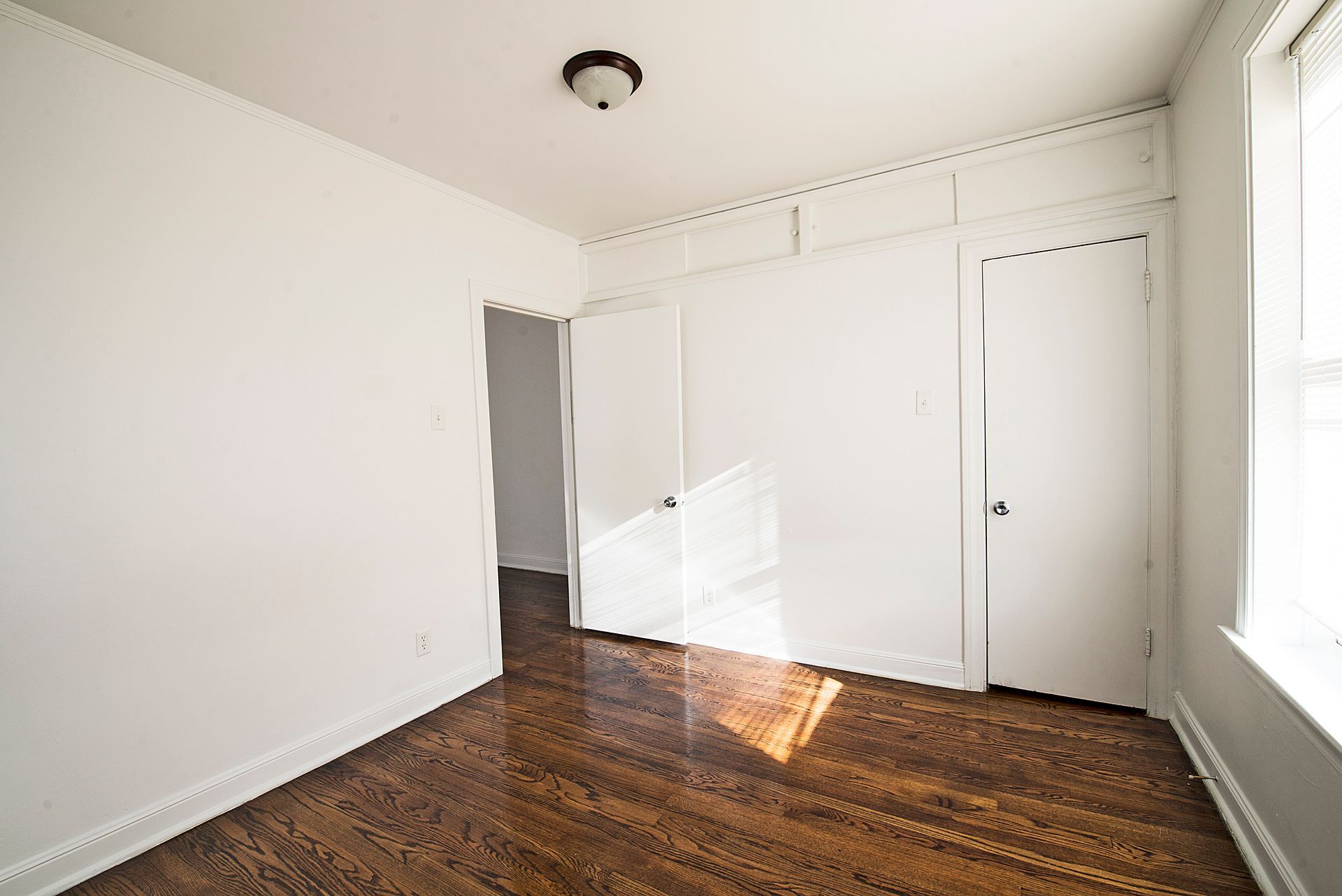 Empty room with white walls, closet doors, and hardwood floors. Sunlight streams through a window.