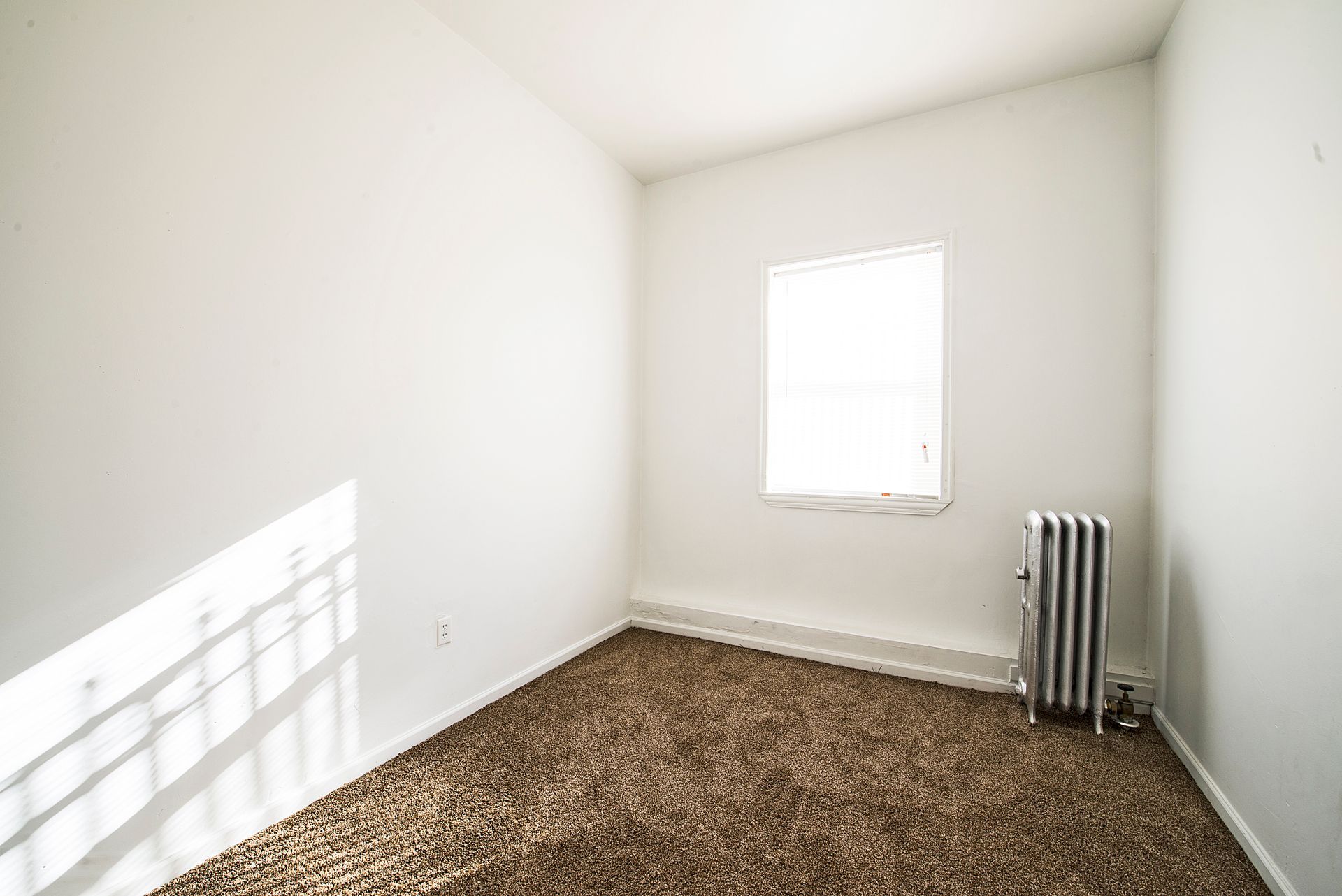 Empty room with brown carpet, white walls, small window, and radiator.
