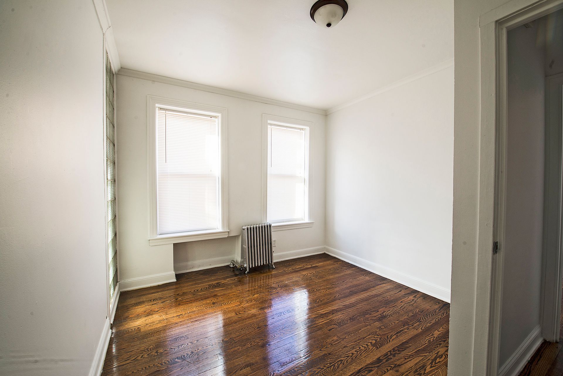 Empty room with hardwood floors, two windows with blinds, and a radiator. White walls and ceiling.