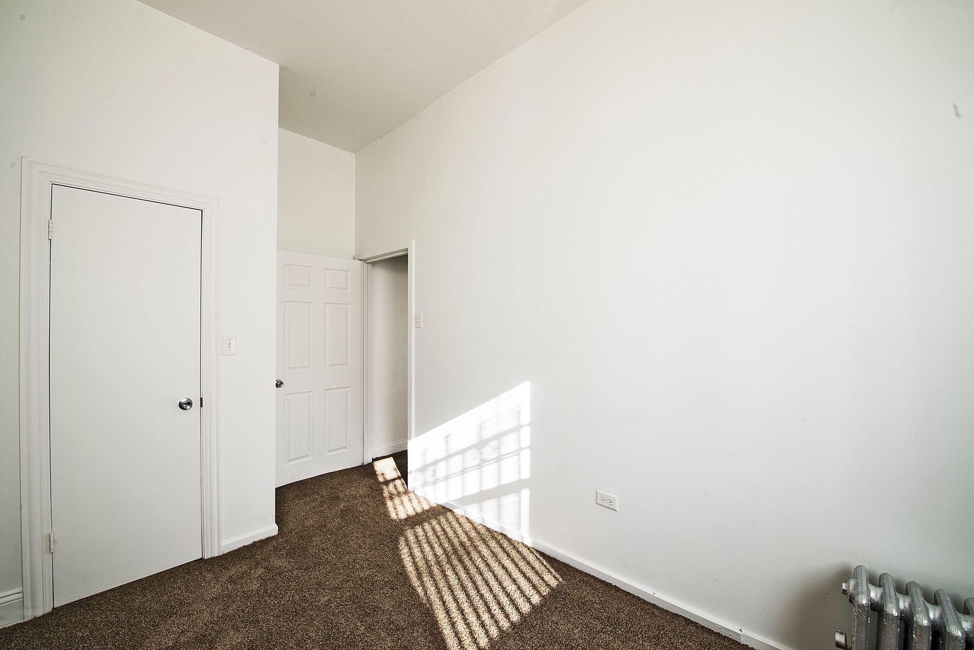 Empty room with white walls, carpeted floor, and closed doors. Sunlight streams through a window.