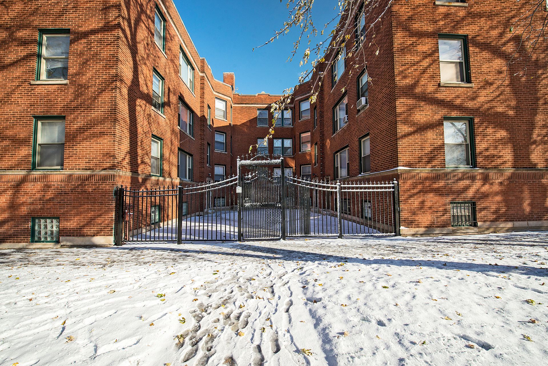 Red brick apartment buildings with snow-covered ground and a fenced walkway in between. Blue sky overhead.
