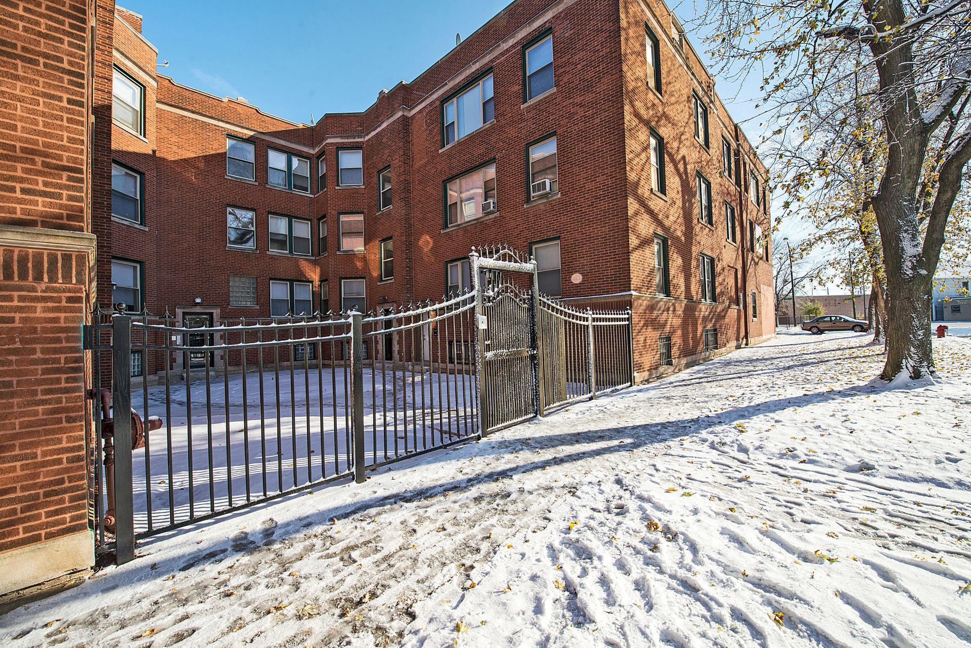 Snowy scene: Red brick apartment building behind a black iron fence.