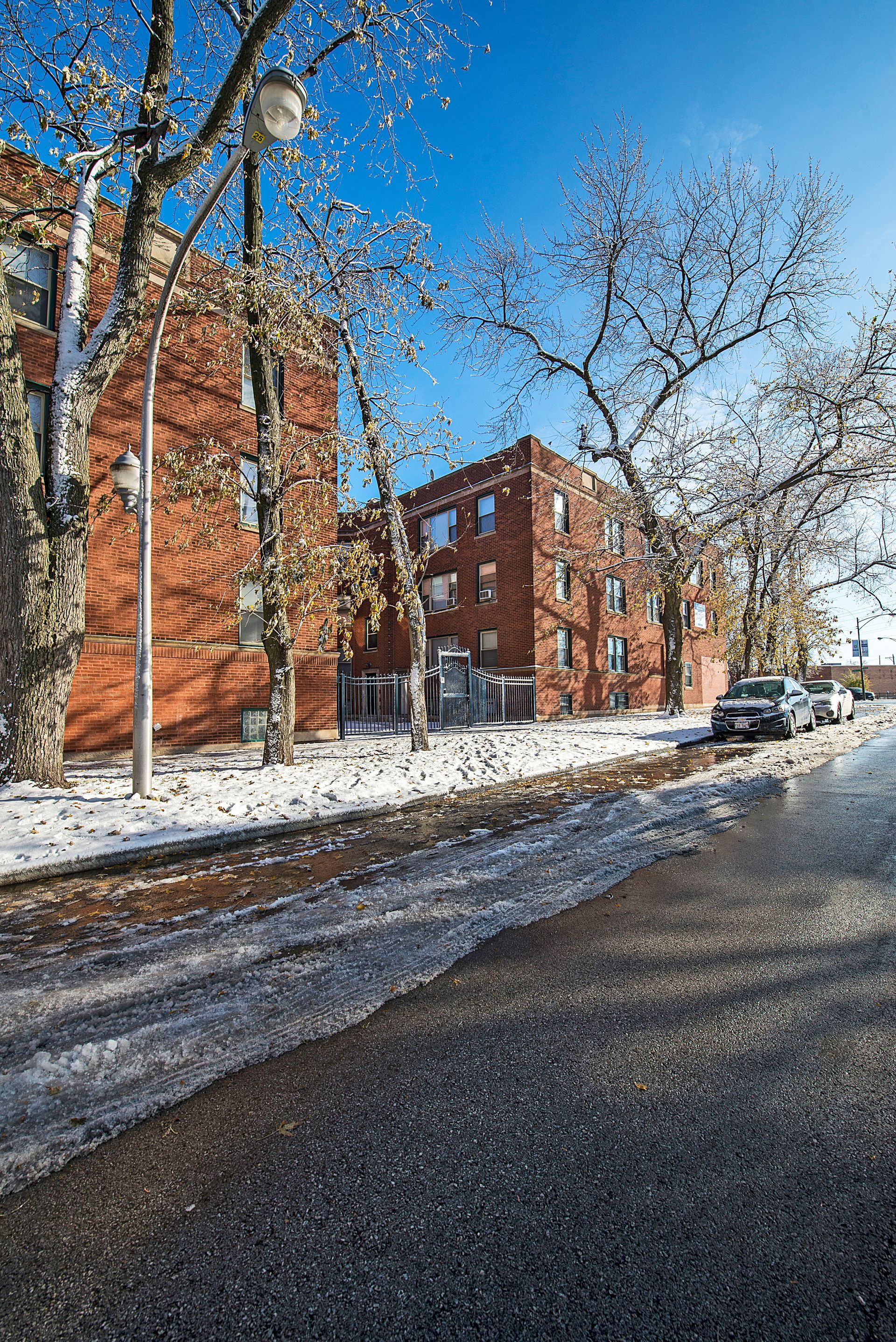 Snow-covered street with red brick buildings and bare trees under a blue sky; cars parked along the curb.