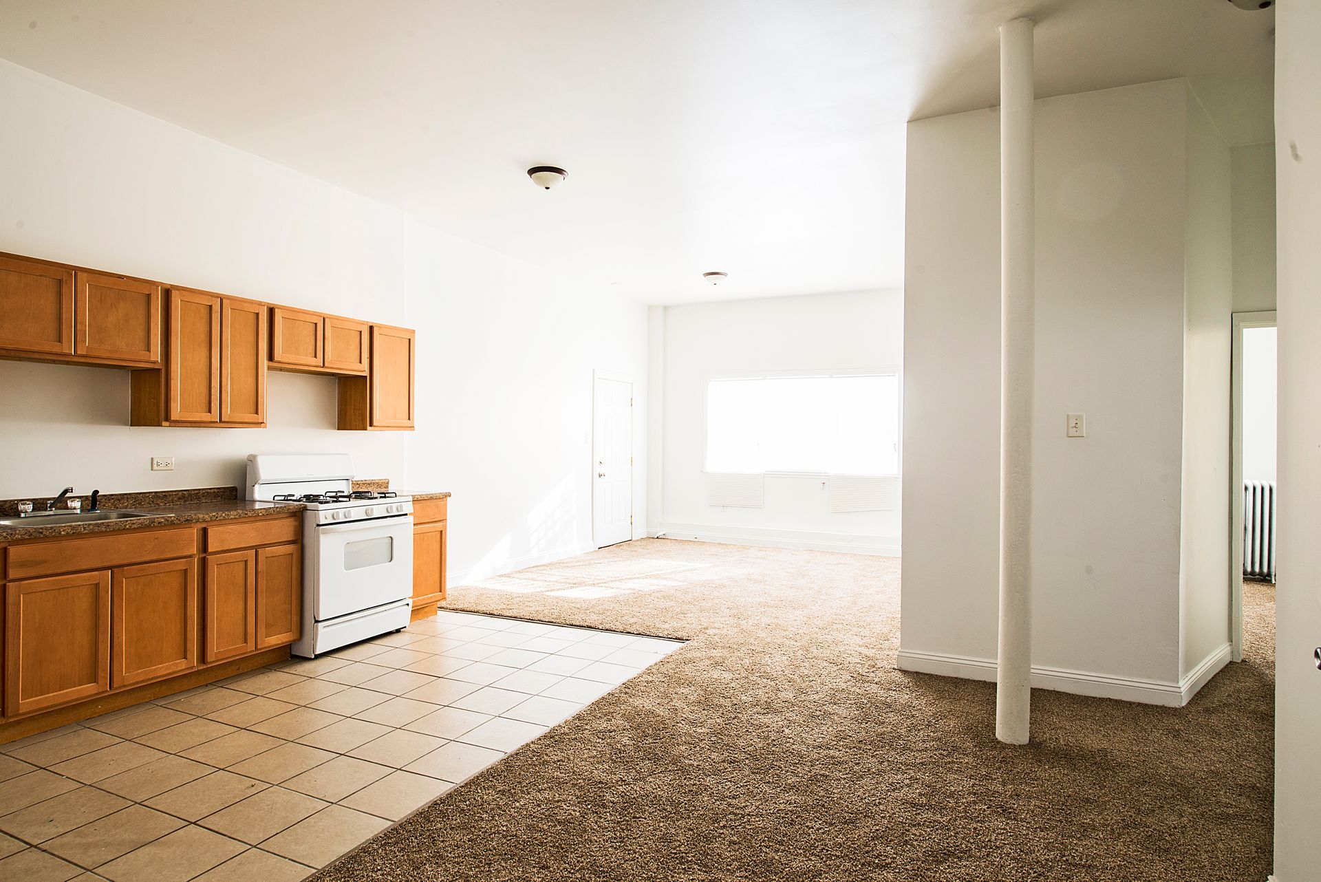 Empty kitchen with wooden cabinets, white stove, and brown carpet.