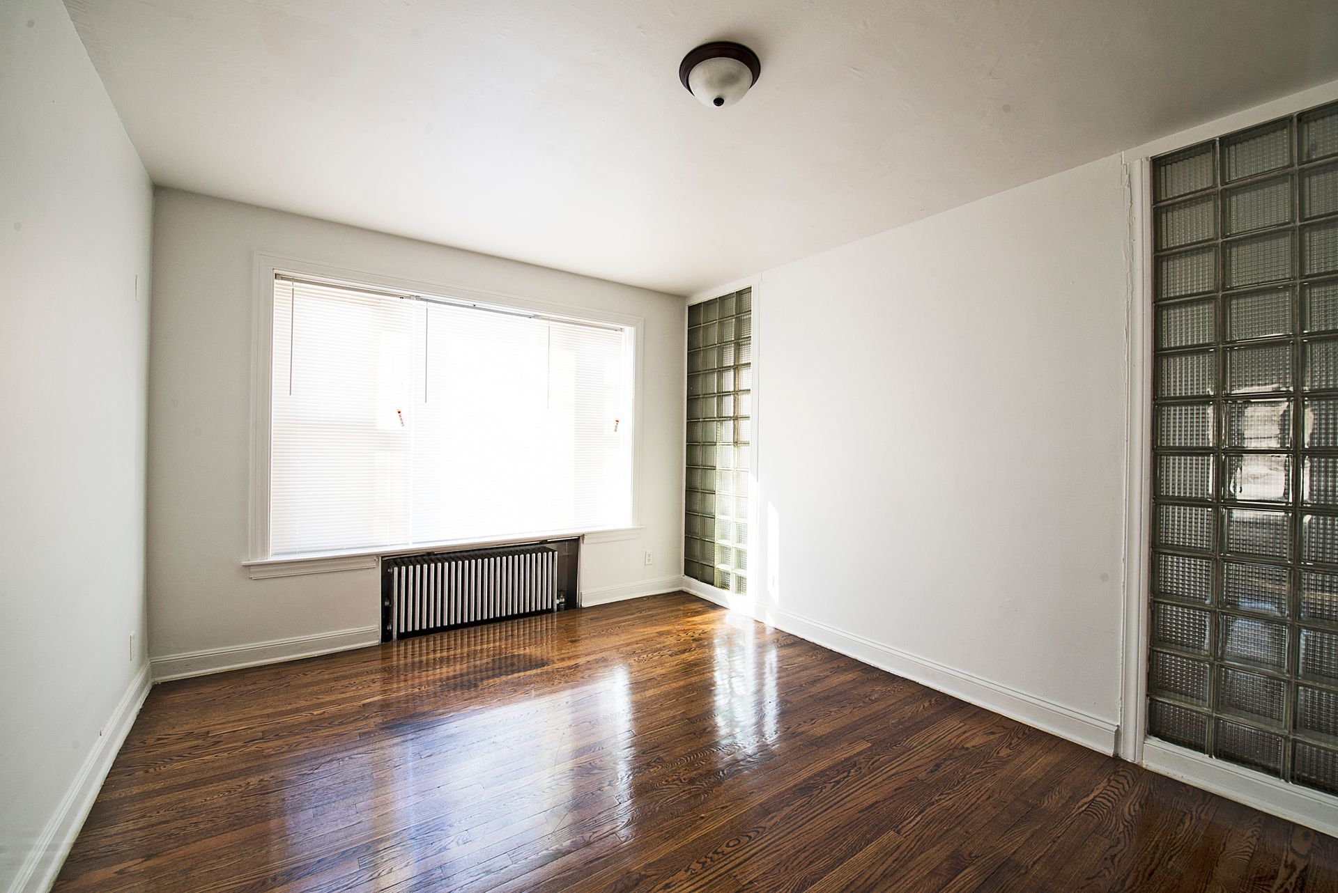 Empty room with hardwood floors, a large window with blinds, and a glass block wall.