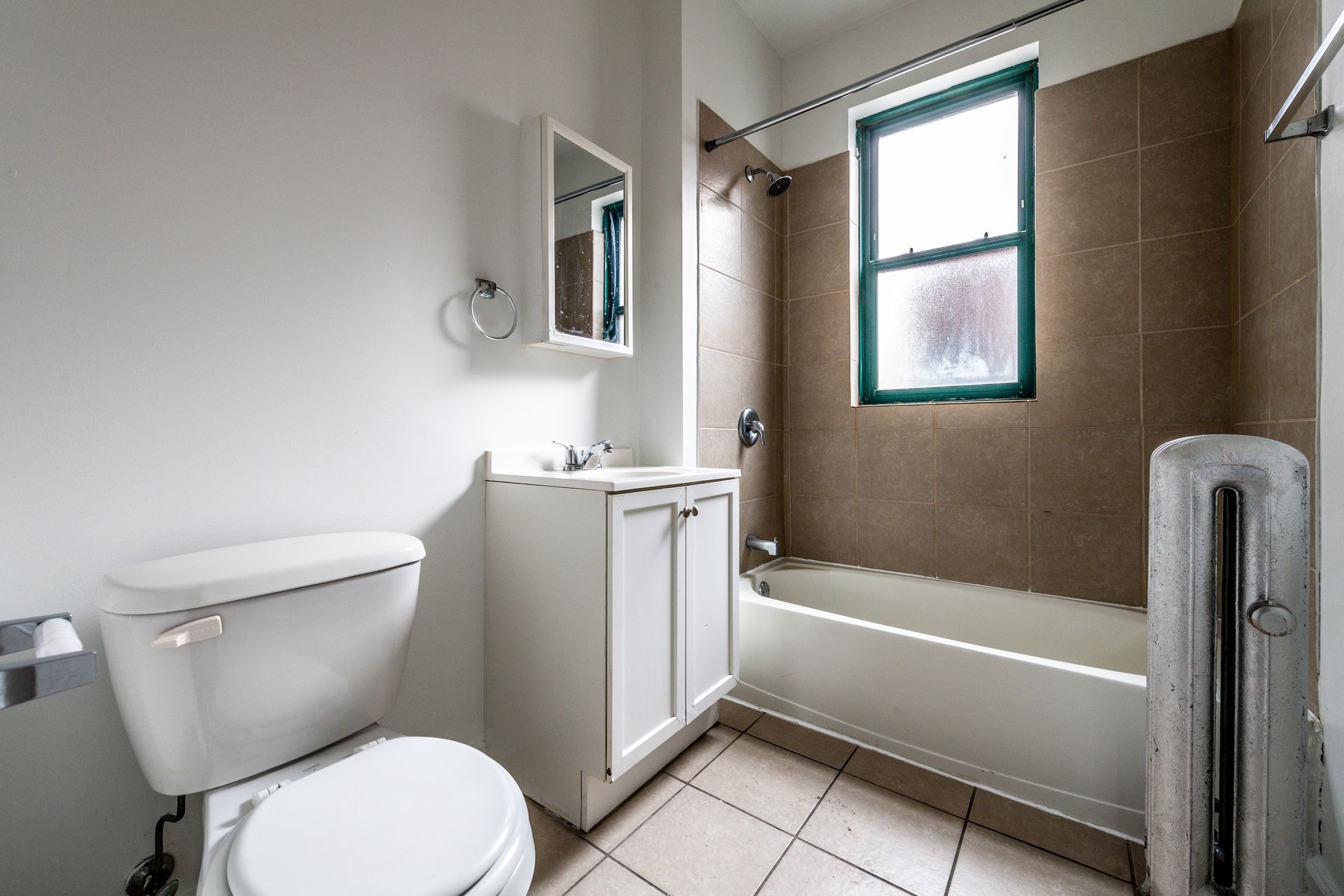 Small bathroom with a toilet, vanity, bathtub, and window with brown tile and white walls.