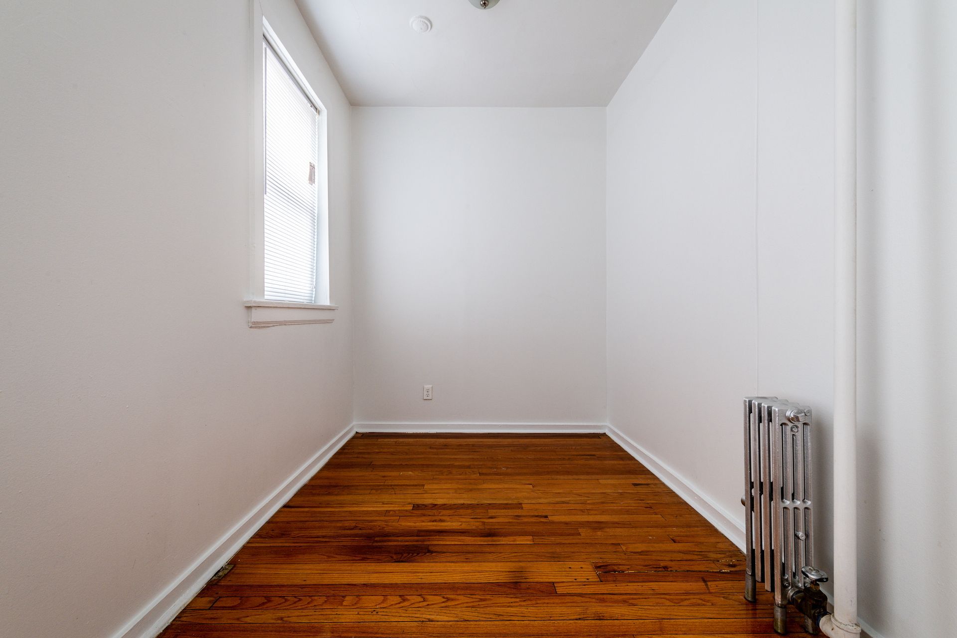 Empty, narrow room with hardwood floor, radiator, and window. White walls and ceiling.