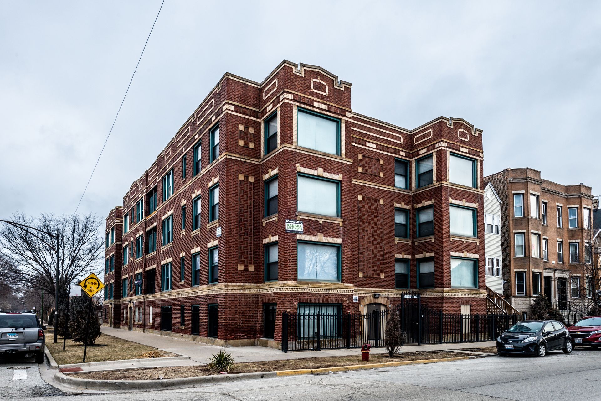 Brick apartment building on a street, overcast day. Cars parked nearby.
