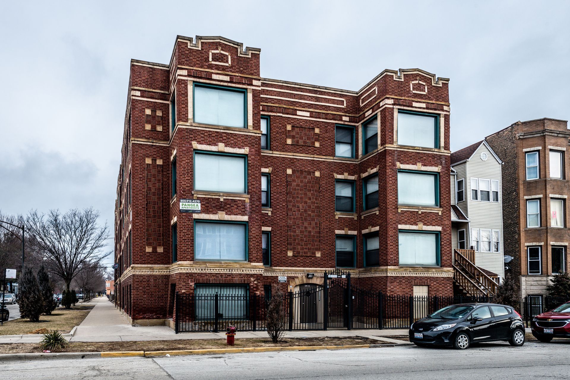 Brick apartment building on a city street, overcast sky. Cars parked in front, sidewalk in view.