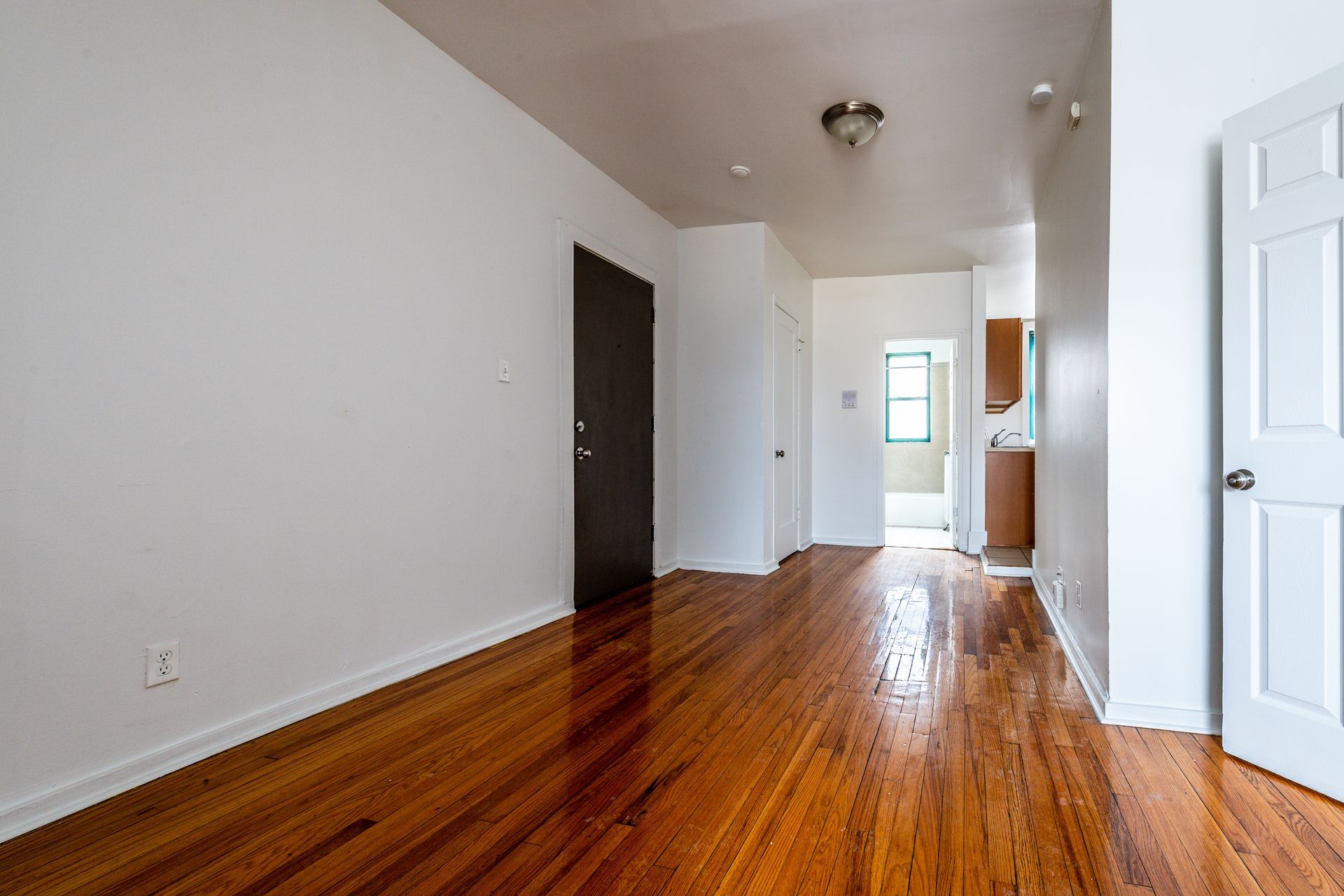Empty apartment interior with wooden floors, white walls, and a hallway leading to a kitchen.