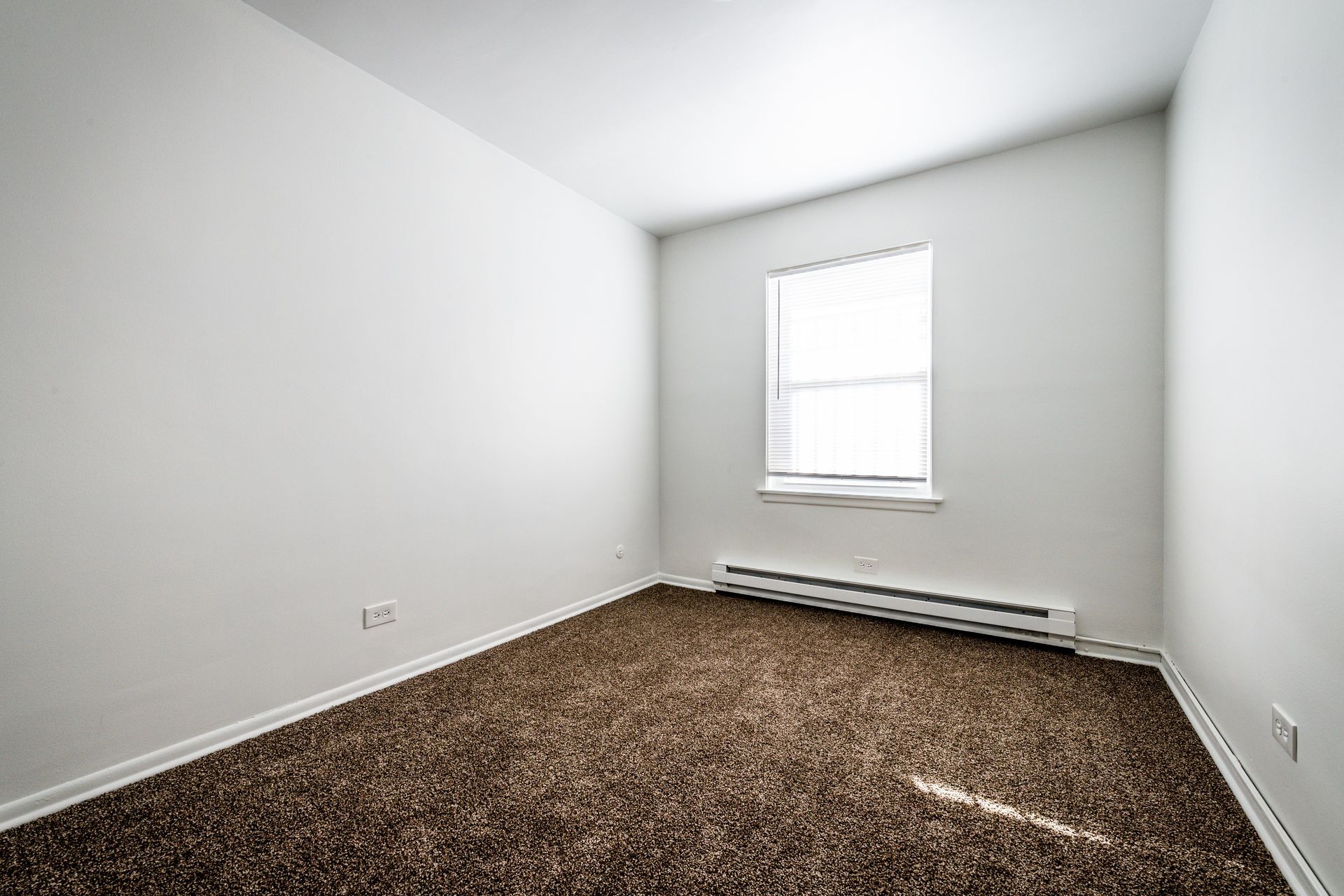 Empty, small room with brown carpet, white walls, and a window with blinds.