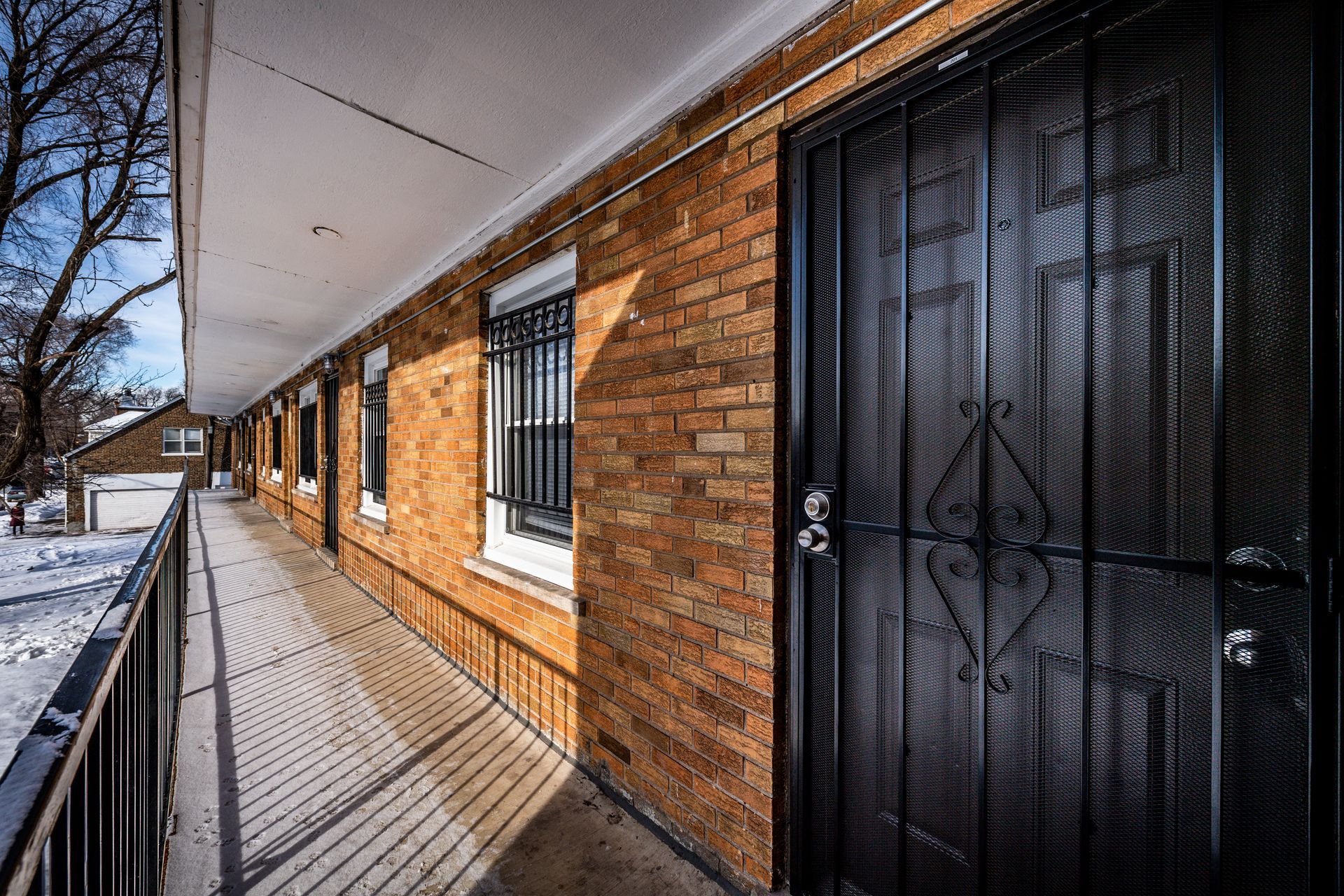 Exterior hallway of a brick building with security doors. Snow on the ground, trees visible in the background.