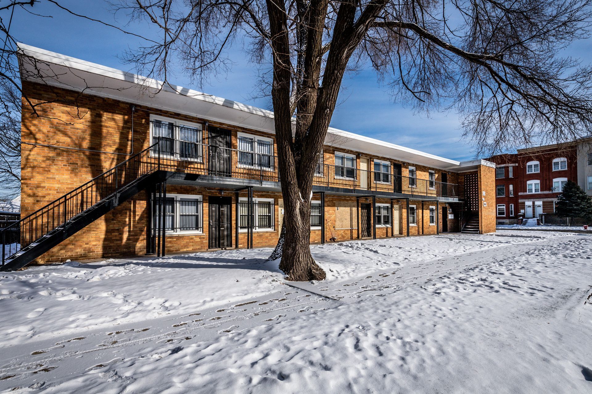 Two-story brick apartment building covered in snow, with bare tree in front.