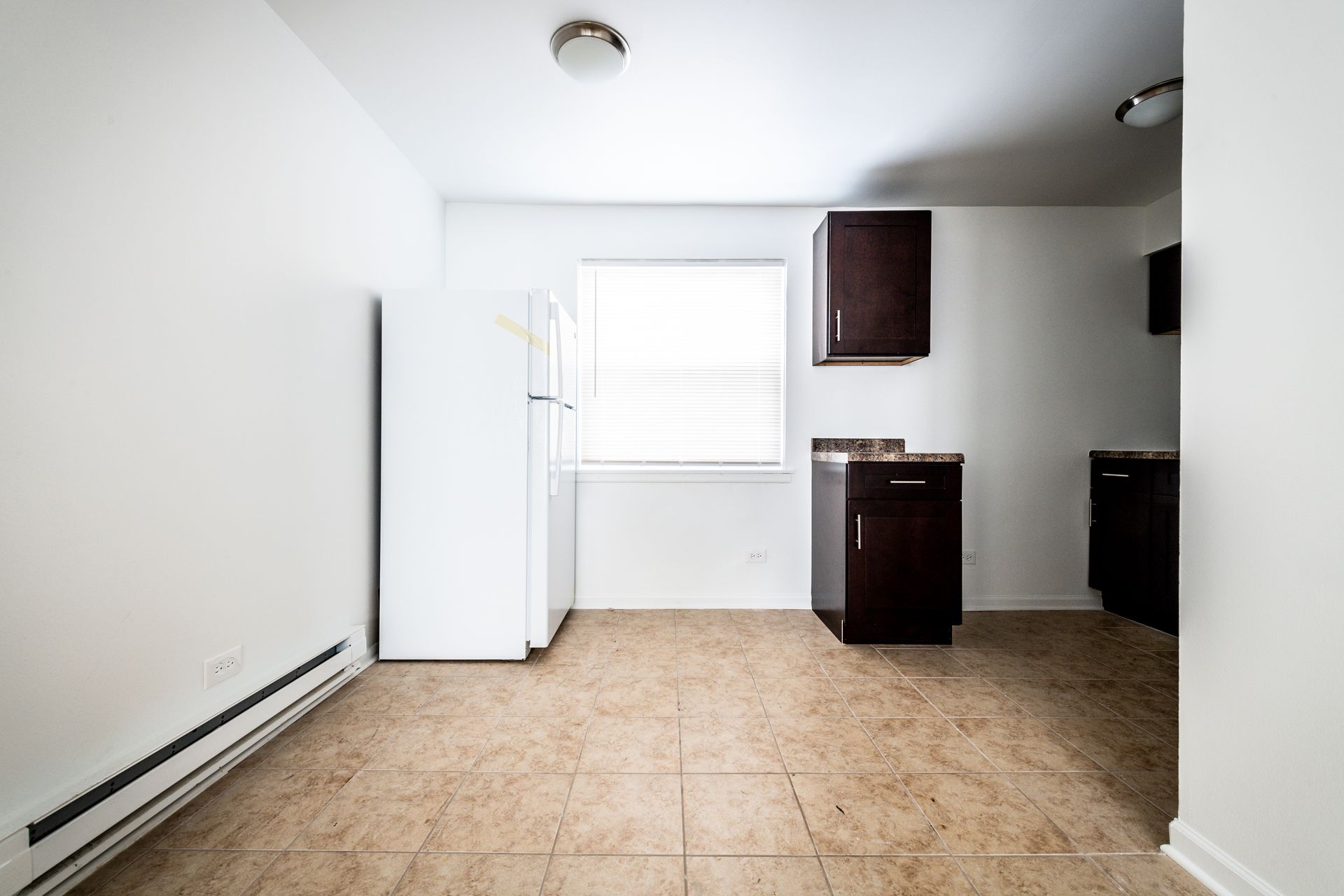 Empty kitchen with white walls, cabinets, fridge, and window. Brown tile floor.