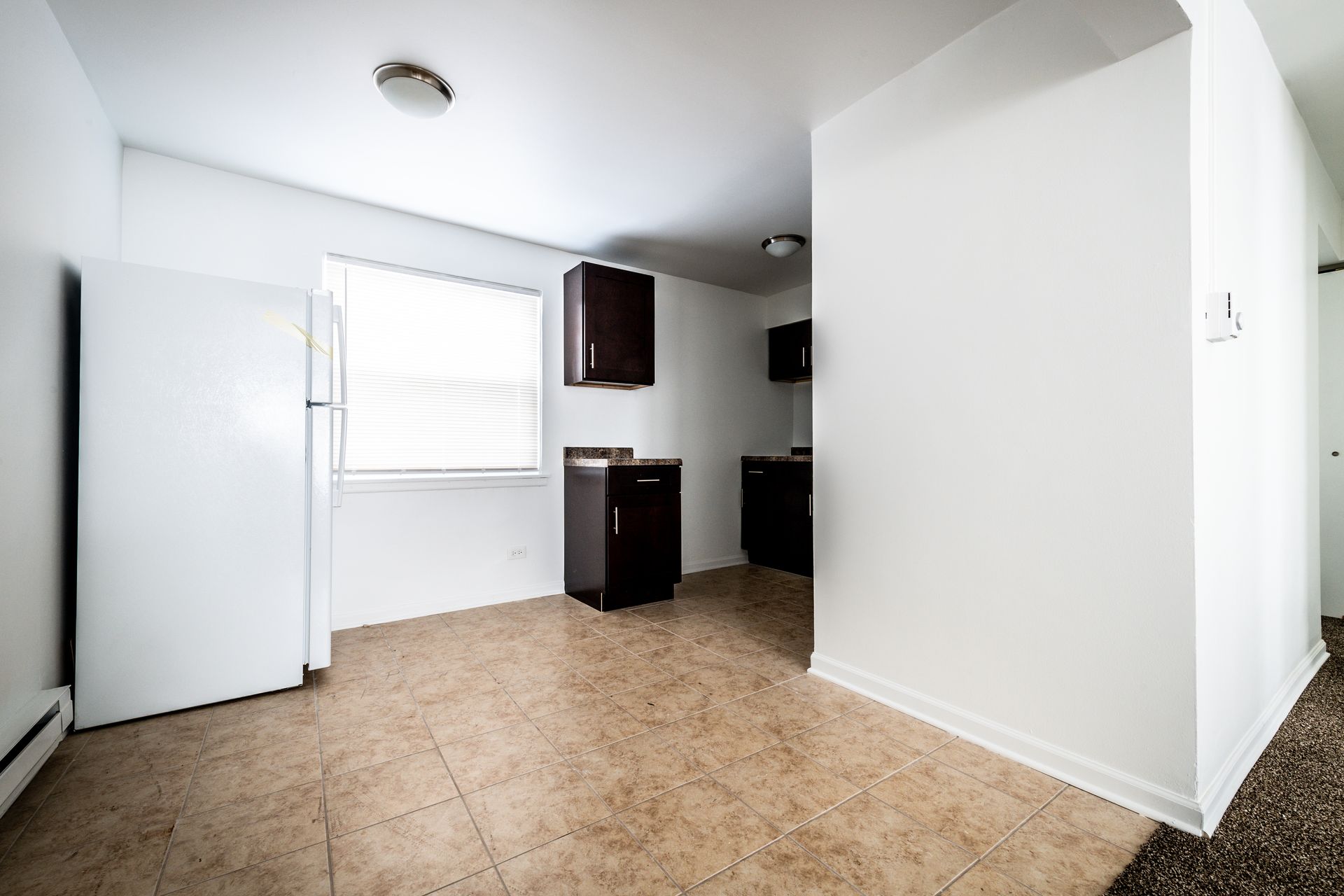 Empty kitchen with white walls, dark cabinets, and tan tiled floor. A window is in the back.