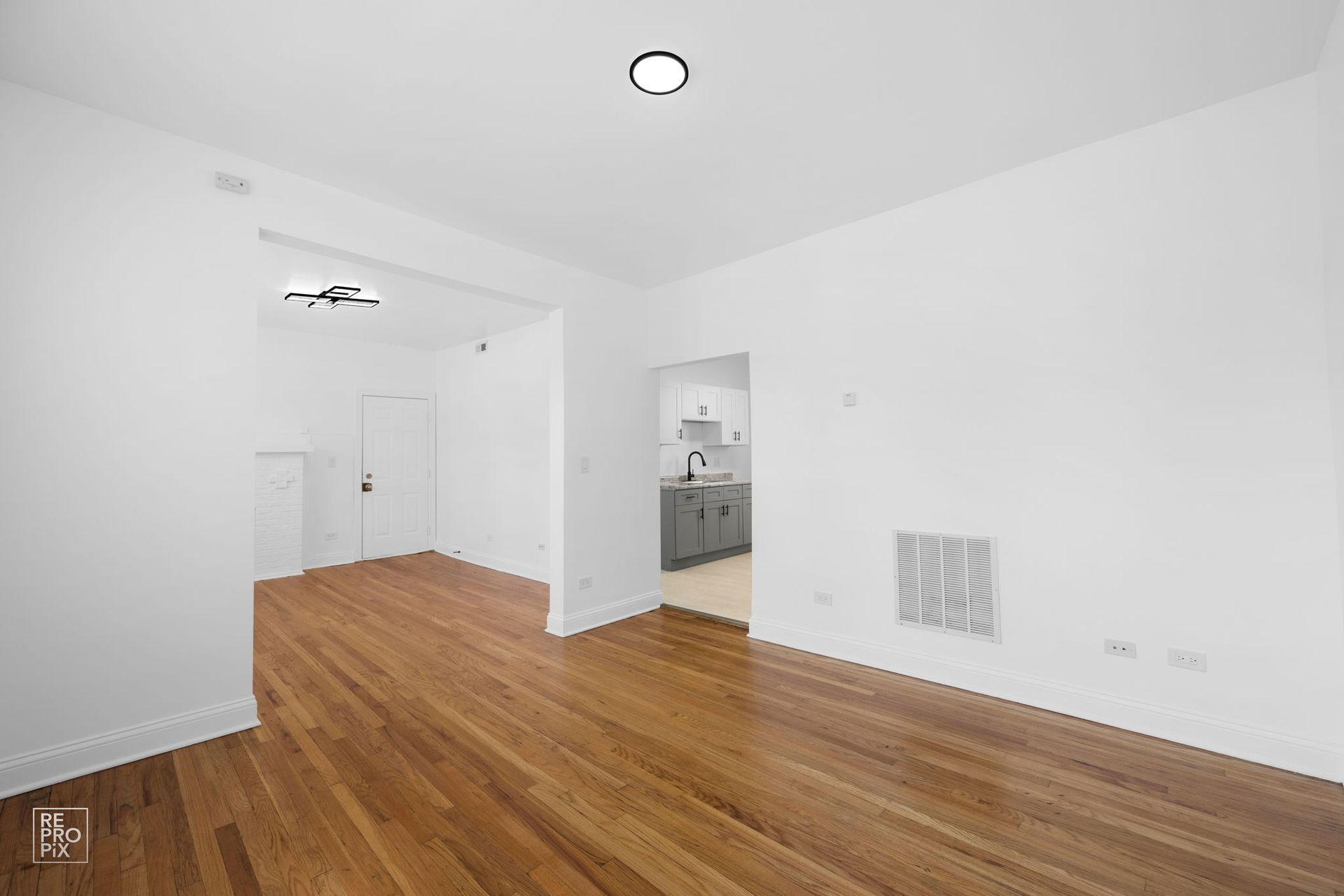 Empty room with hardwood floors, white walls, and an open doorway leading to a kitchen.