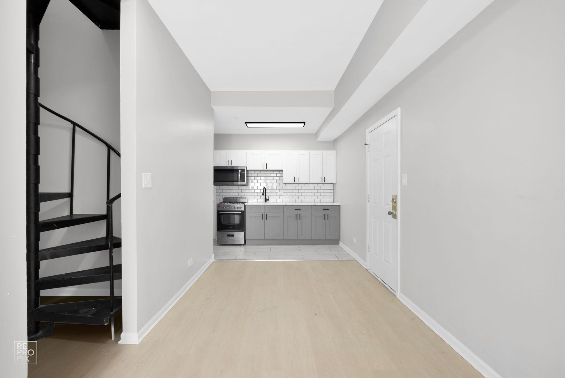 Interior shot of a small apartment, featuring a spiral staircase, kitchen, and a door, with light-colored walls and flooring.