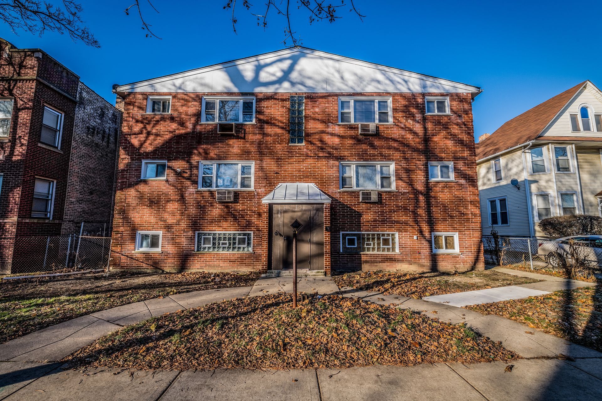 Two-story brick apartment building on a sunny day; fallen leaves on the ground.