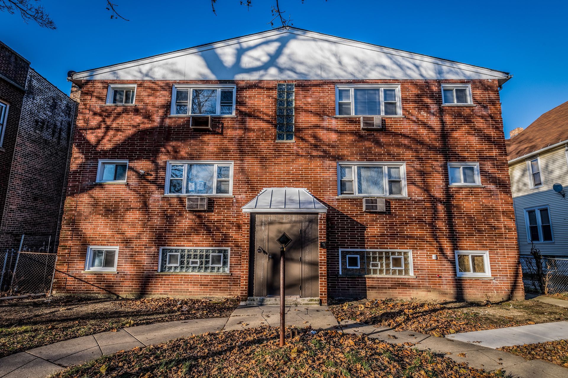 Brick multi-unit building with multiple windows under a blue sky, front entrance centered, fallen leaves.