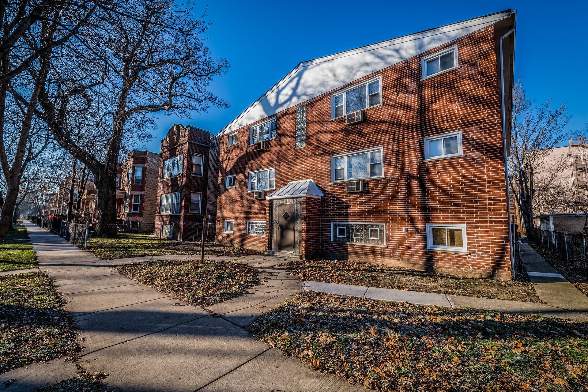 Brick apartment building with sidewalk and bare trees on a sunny day. Leaves cover the ground.