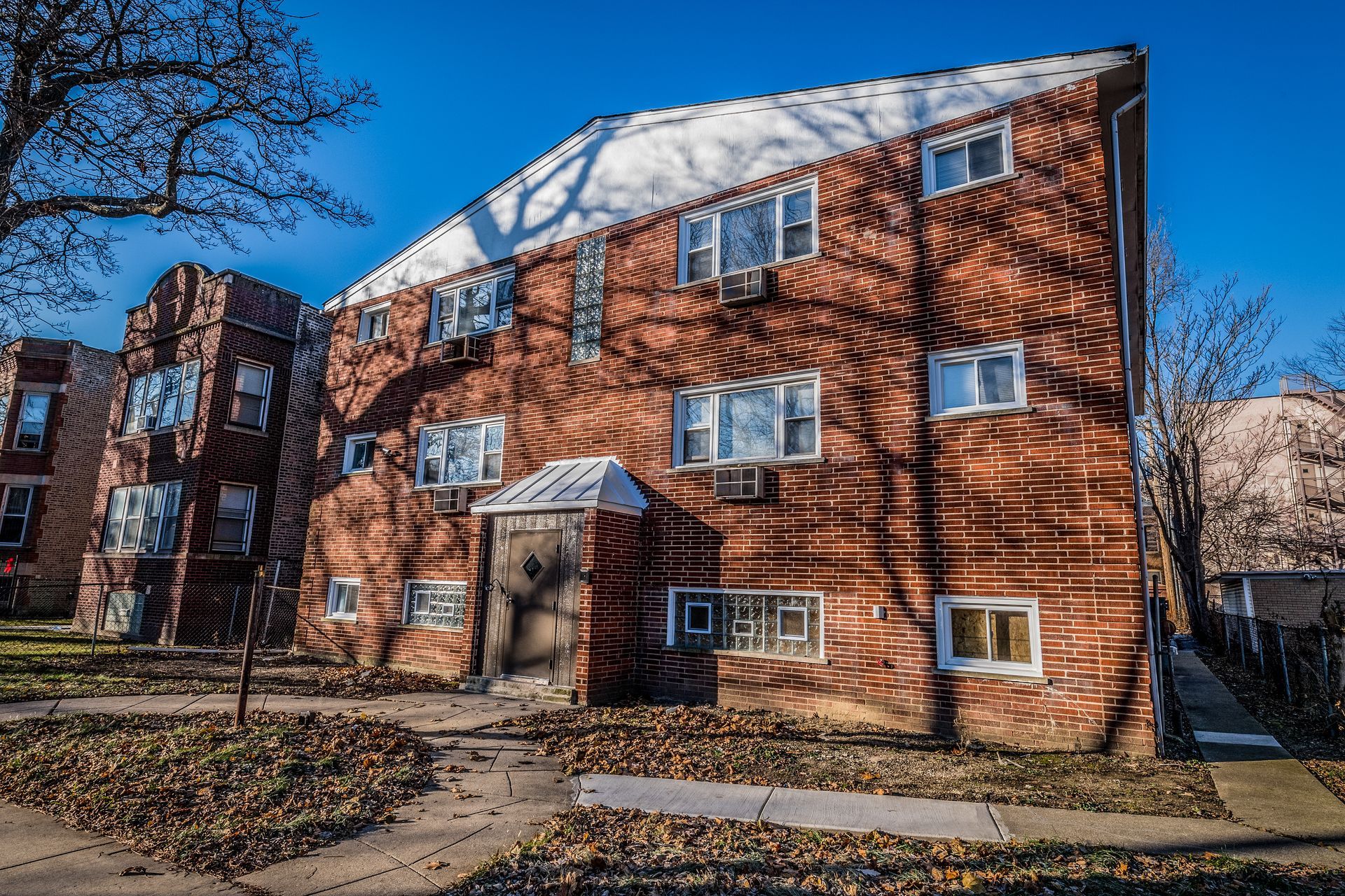 Brick apartment building with rectangular windows and a snow-covered roof, casting tree shadows on the facade.