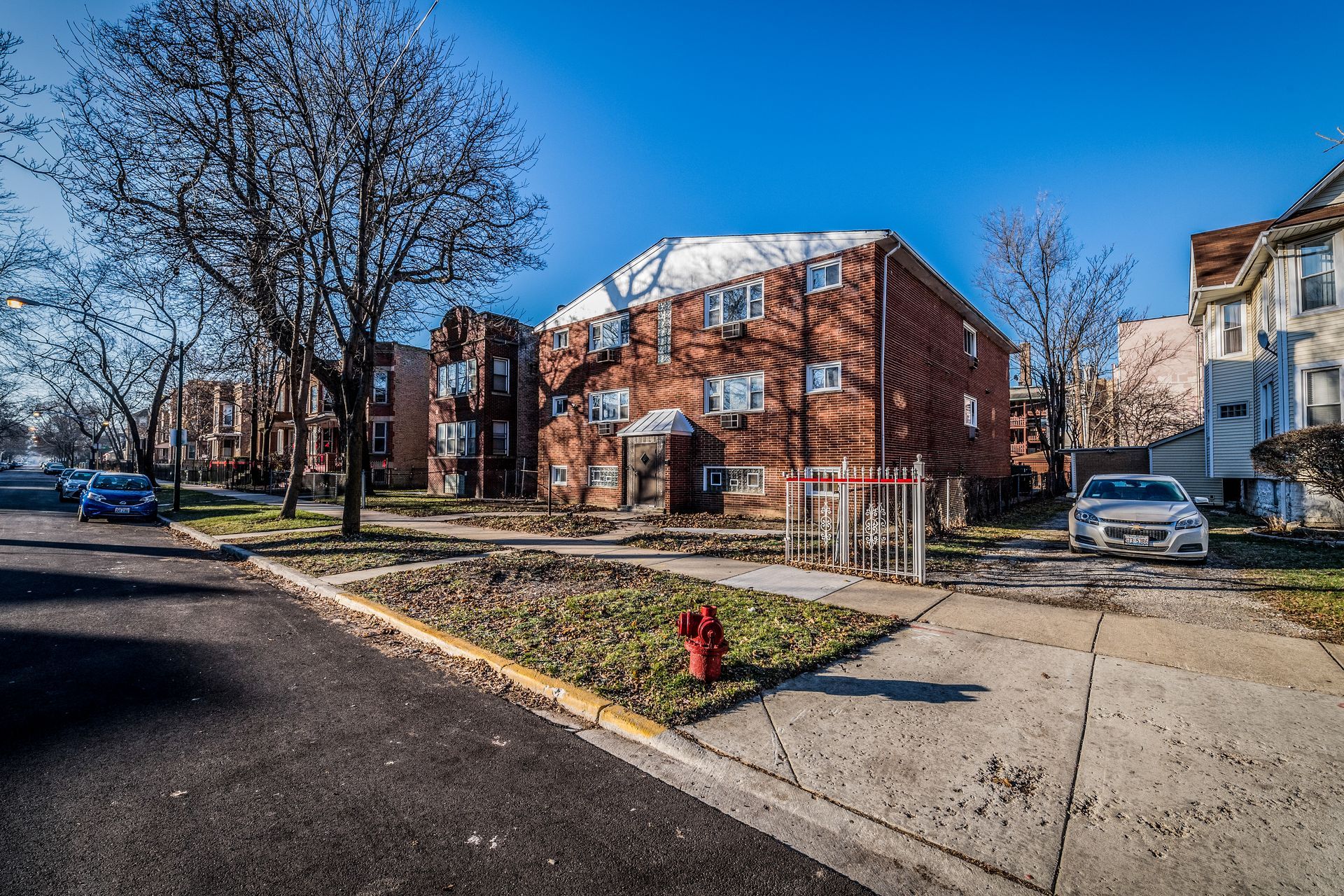 Brick apartment building with bare trees on a city street; parked cars and clear blue sky.