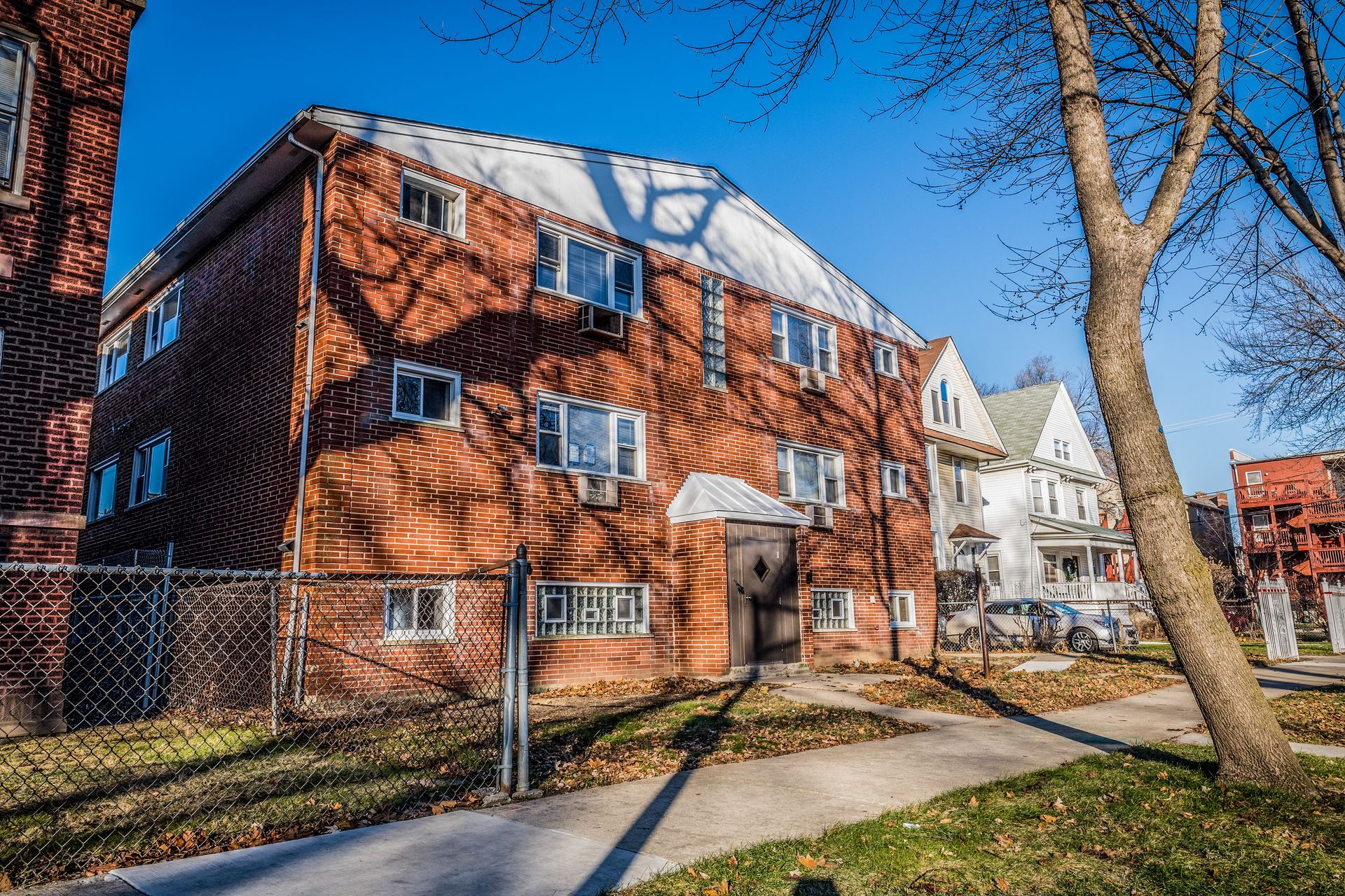 Brick apartment building on a sunny day with tree shadows. Sidewalk and fenced yard.