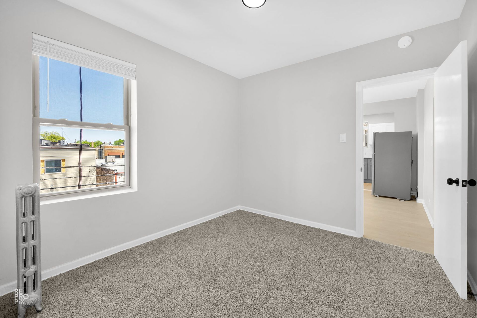 Empty room with gray carpet, a window, and a doorway to a kitchen with a refrigerator.
