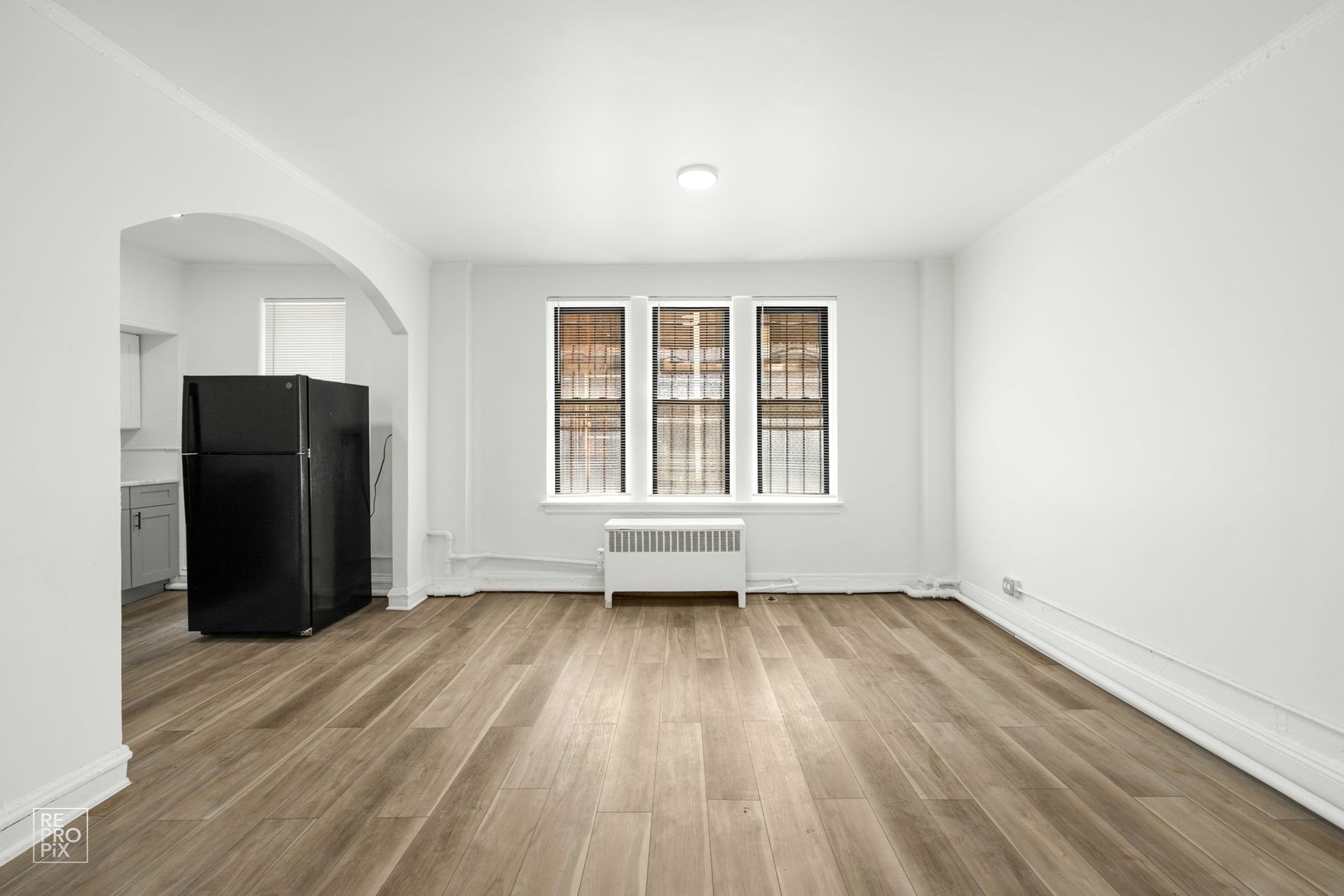 Empty apartment interior, with hardwood floors, white walls, and a black refrigerator.